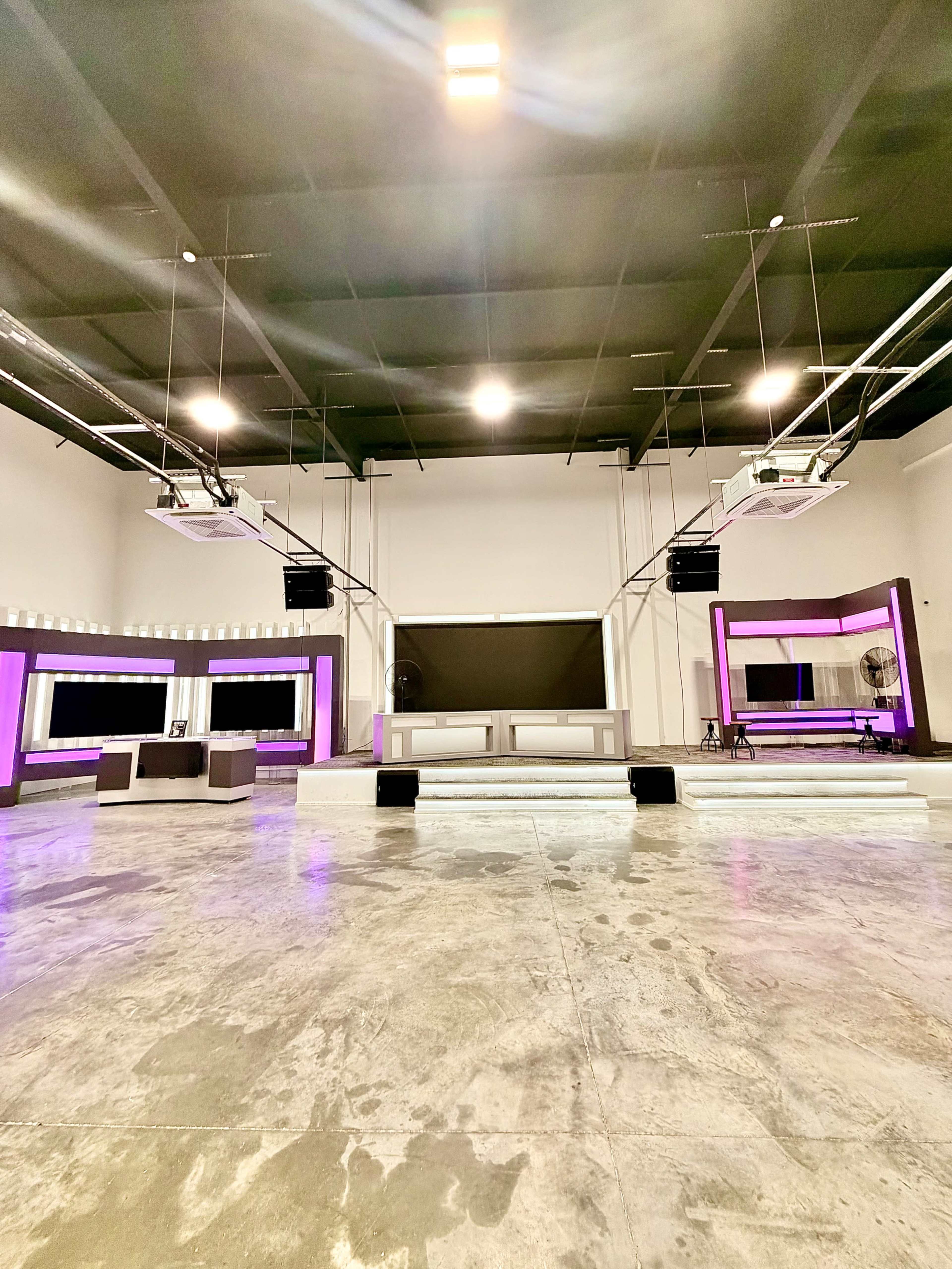 The image shows a spacious studio interior with white walls, a polished concrete floor, and modern lighting, featuring large mirrors and purple accents throughout.
