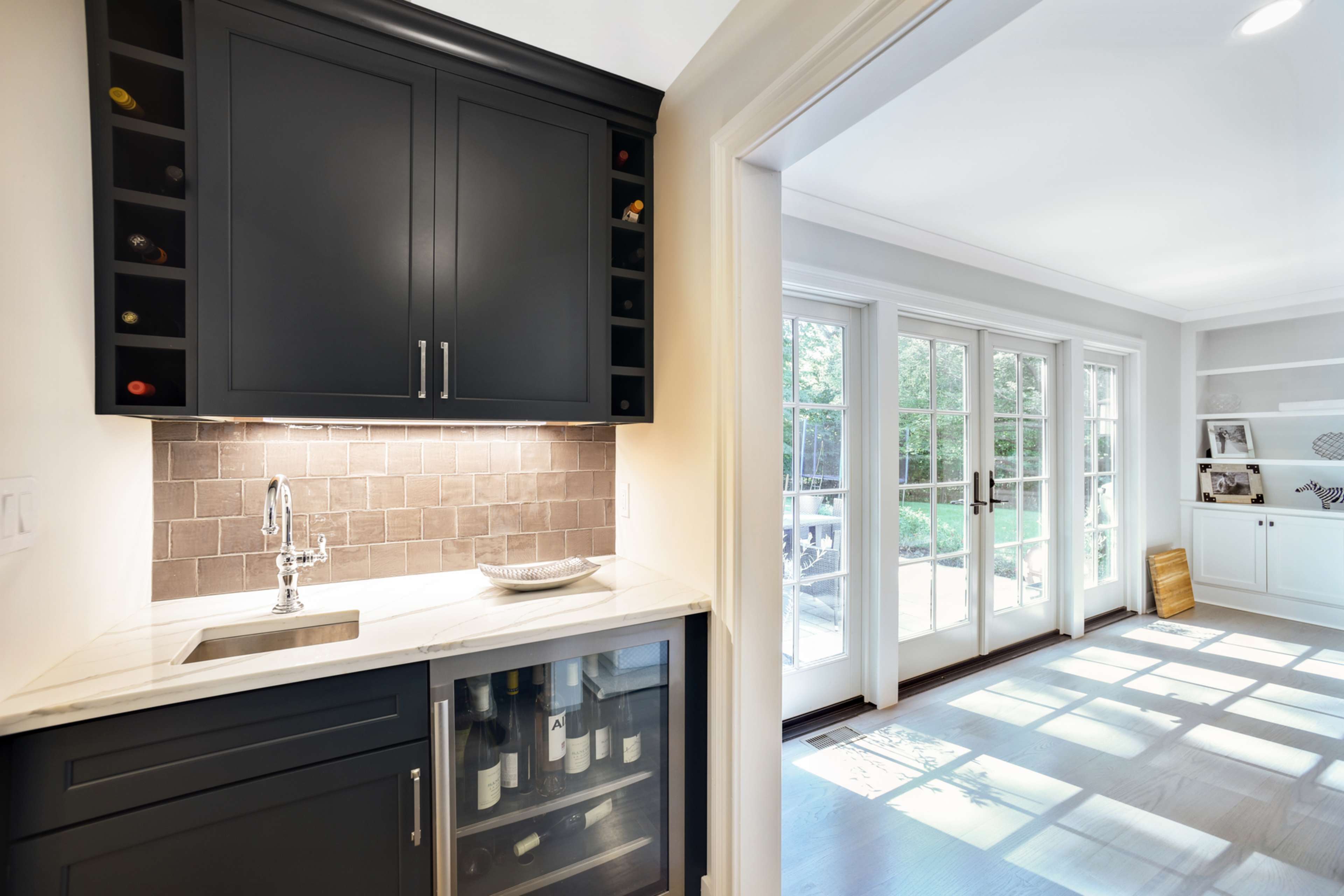 A dark cabinetry wine bar features a marble countertop, sink, and wine fridge, leading to a sunny room with French doors.