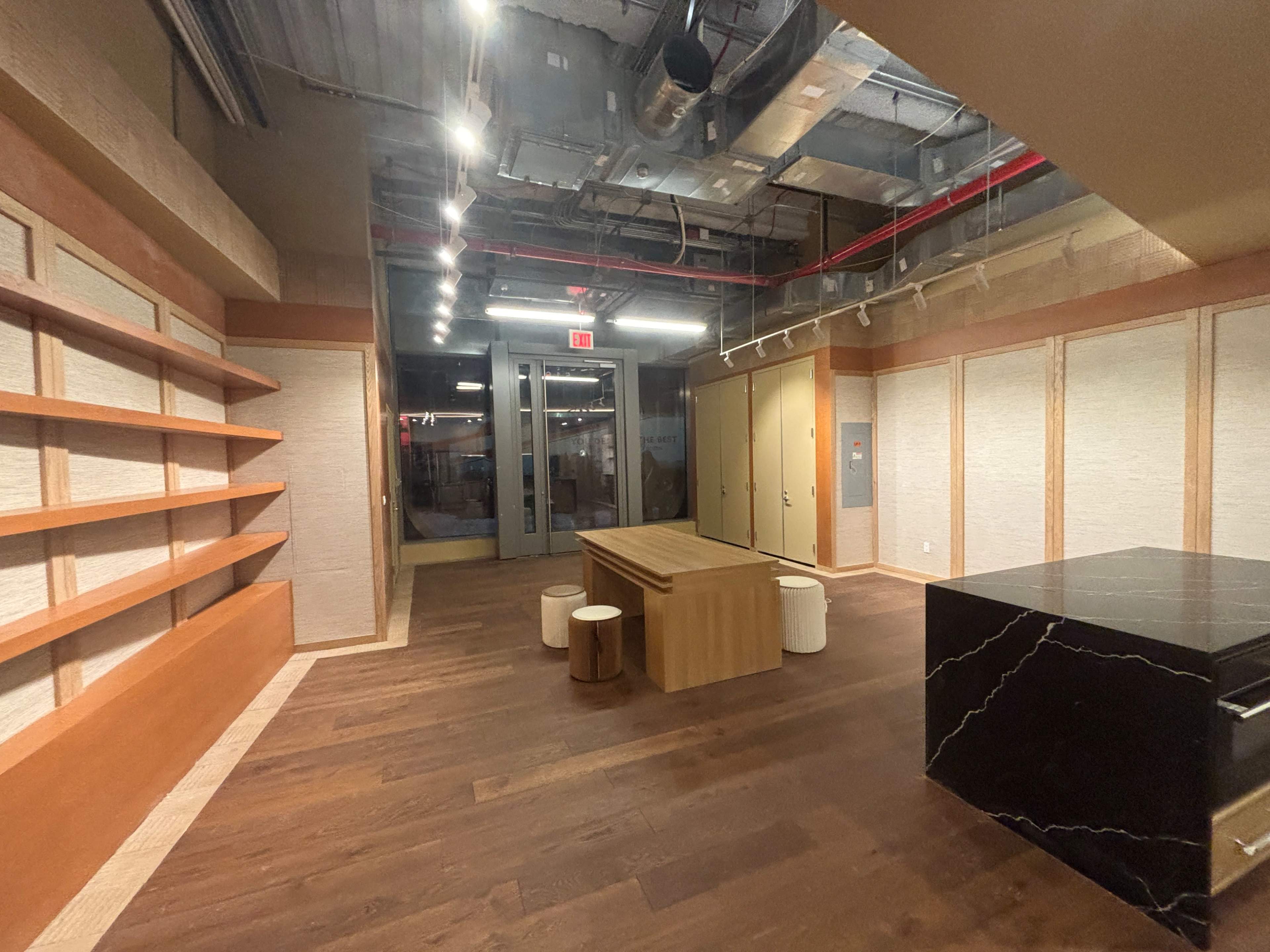 The image shows a spacious interior with wooden floors, a large table, shelves on one wall, and a black marble countertop in a modern setting.