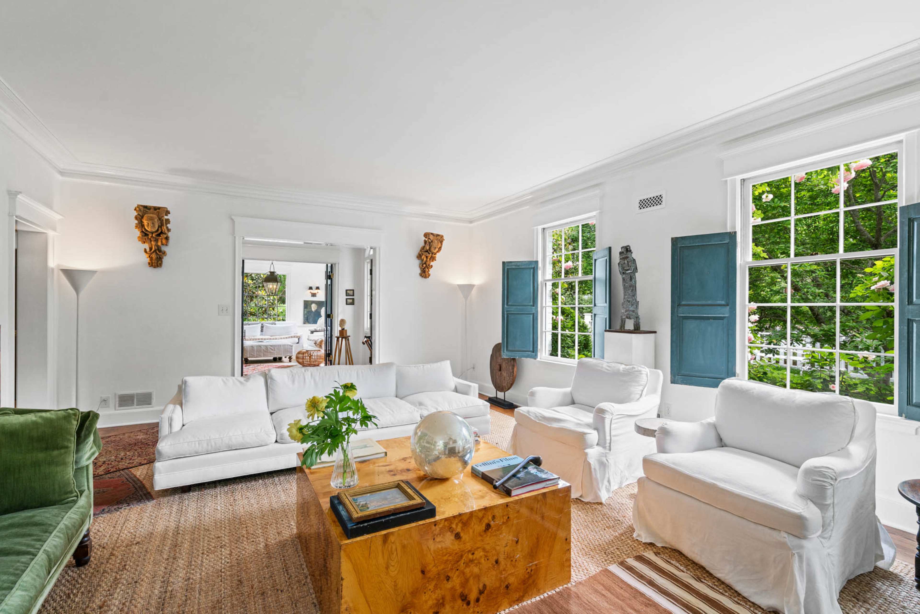 The image shows a bright living room featuring white sofas, a wooden coffee table, and large windows with green shutters, complemented by decorative sculptures and plants.