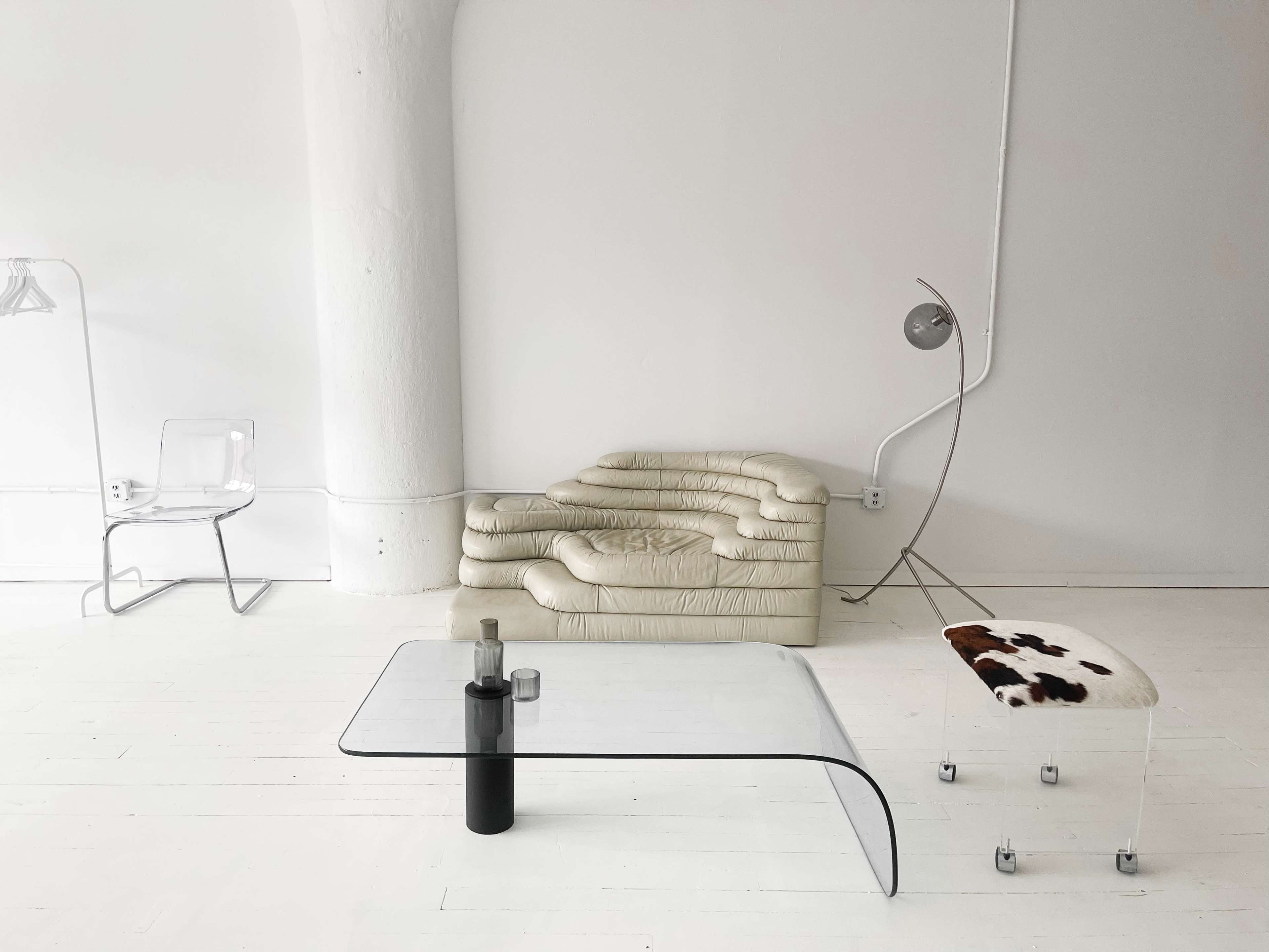 A minimalist interior with a clear glass coffee table, a curved beige sofa, a transparent chair, and a unique bench with a cow print design.