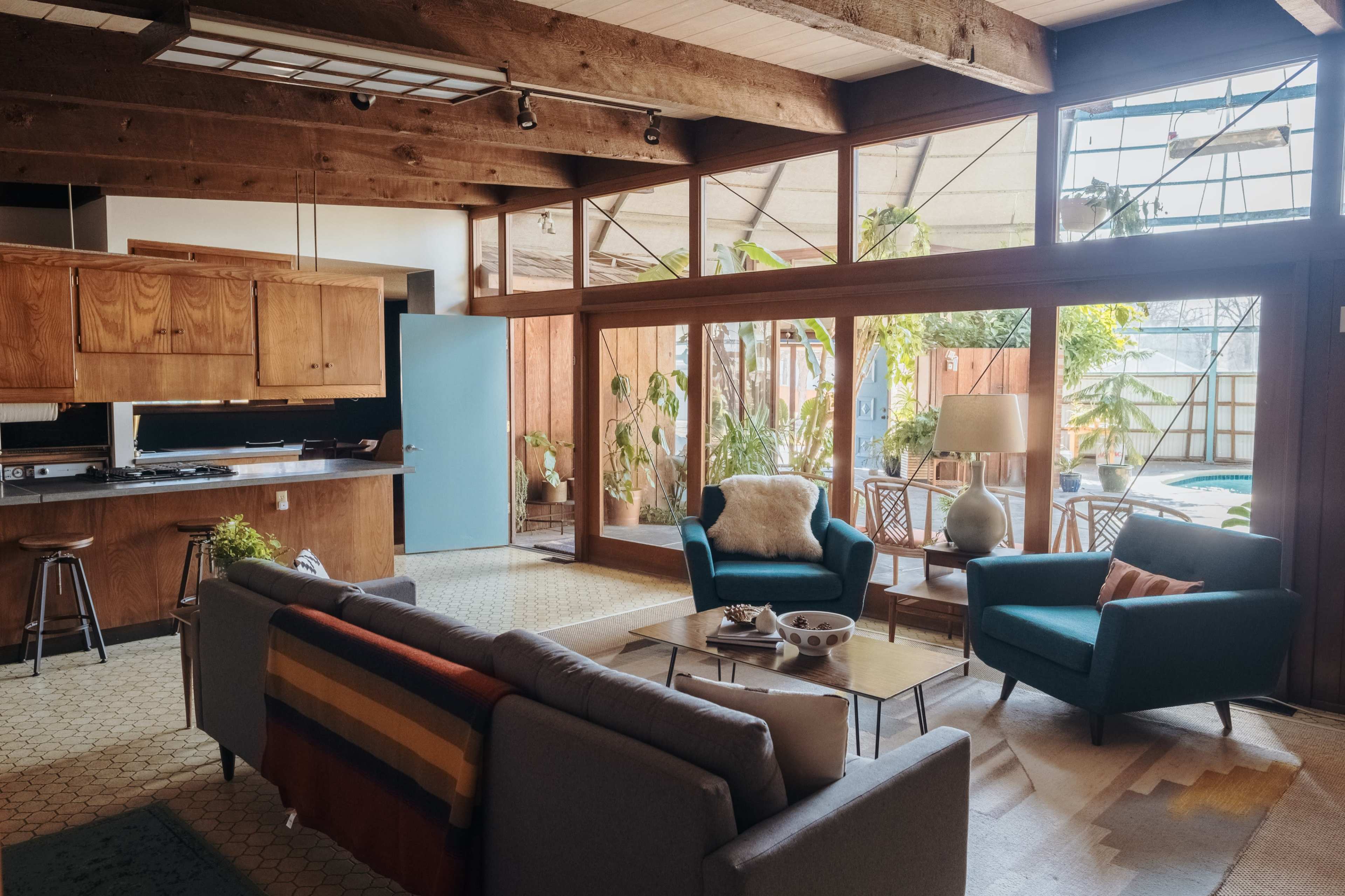 The image shows a spacious living room with large windows, modern furniture, and an open kitchen area visible in the background.