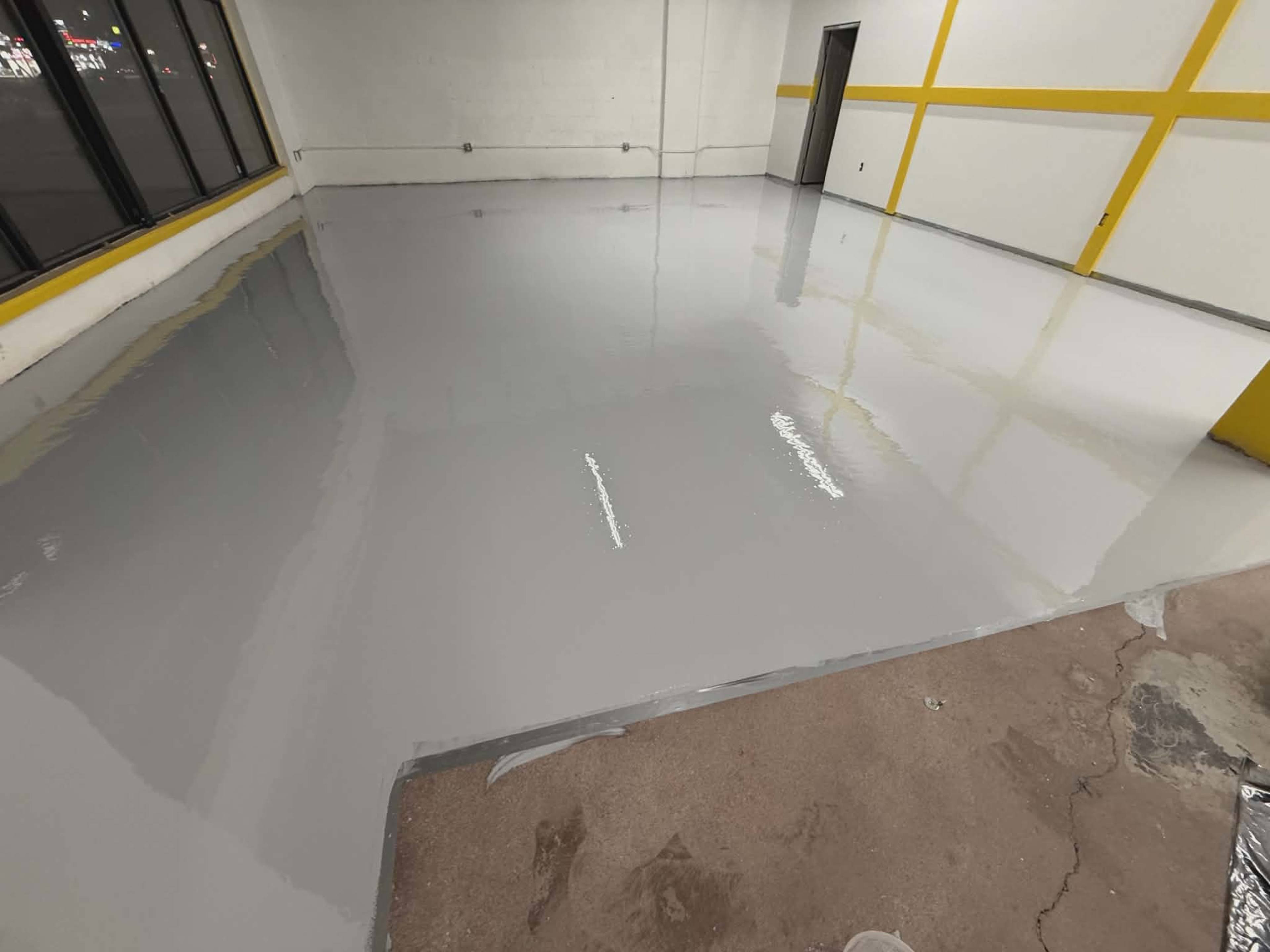 The image shows a clean, freshly coated gray epoxy floor in an empty room with yellow accent stripes on the walls and large windows.