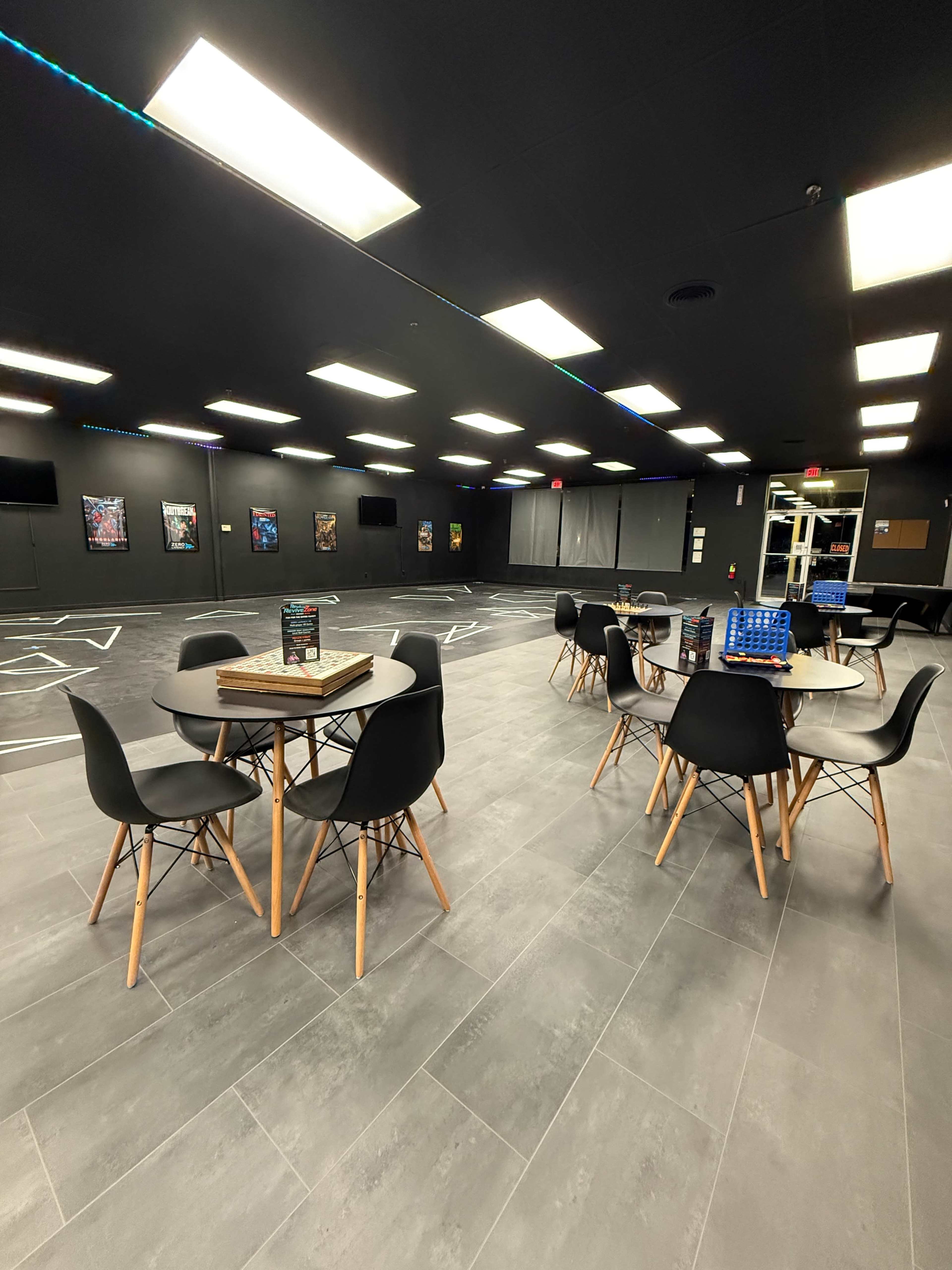 The image shows a spacious, well-lit room with several round tables and chairs arranged on a tiled floor, featuring artwork on the walls and a large unobstructed area in the background.