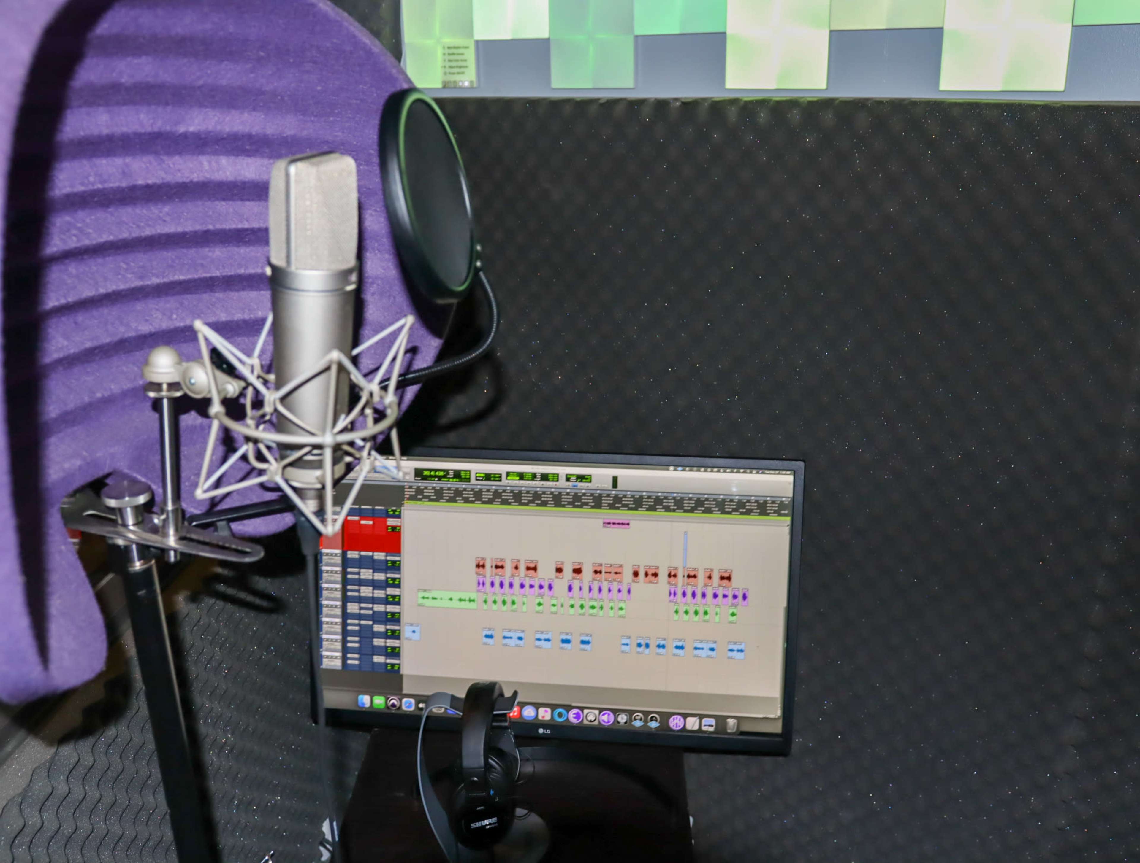 The image shows a microphone with a pop filter next to a computer monitor displaying a music production software interface, all set within a soundproof recording booth.