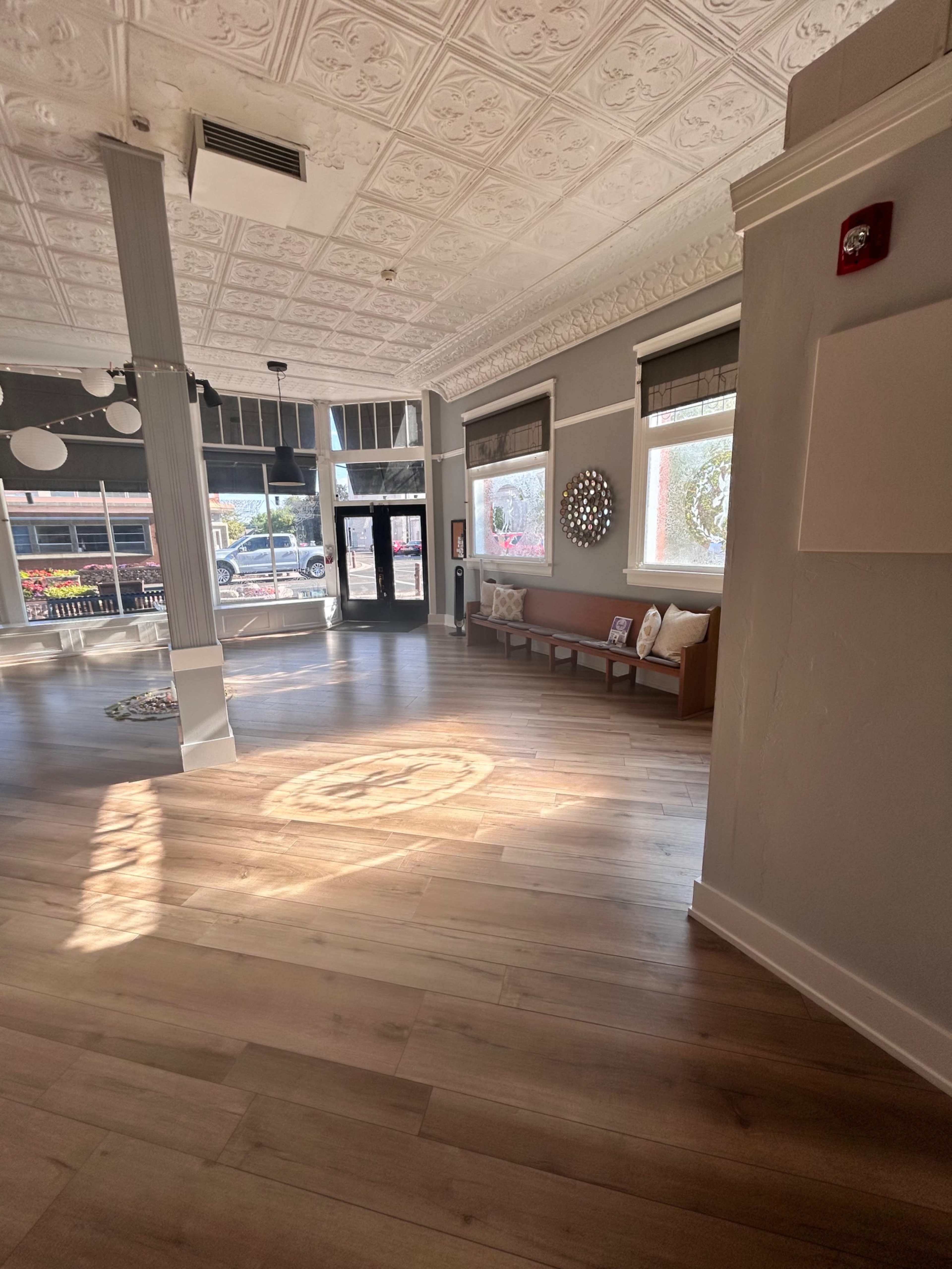 The image shows a well-lit interior with hardwood flooring, featuring a seating area, decorative wall art, and large windows.