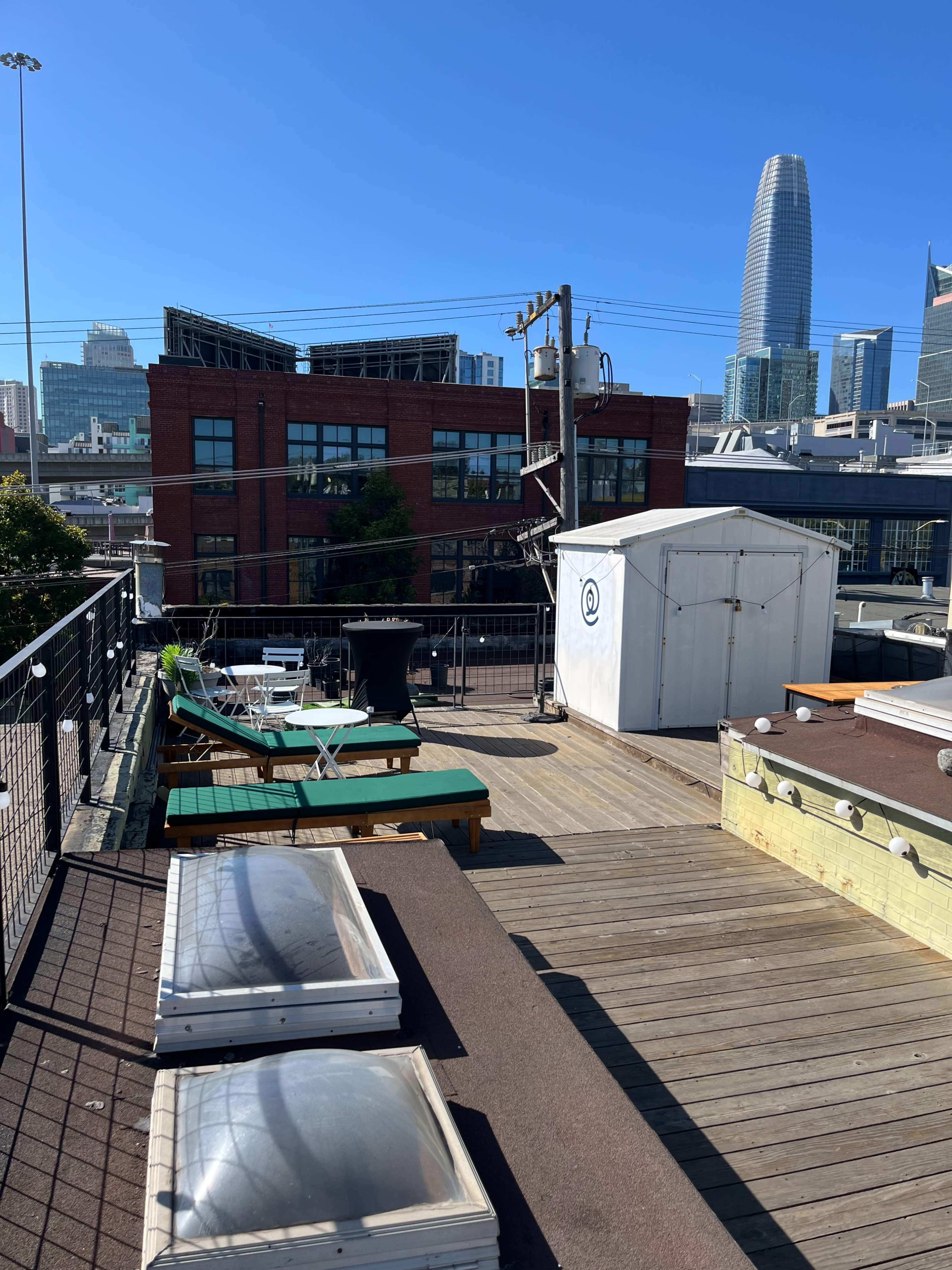 The image shows a rooftop space featuring wooden decking, lounge chairs, and solar panels, with a skyline view of modern buildings in the background.