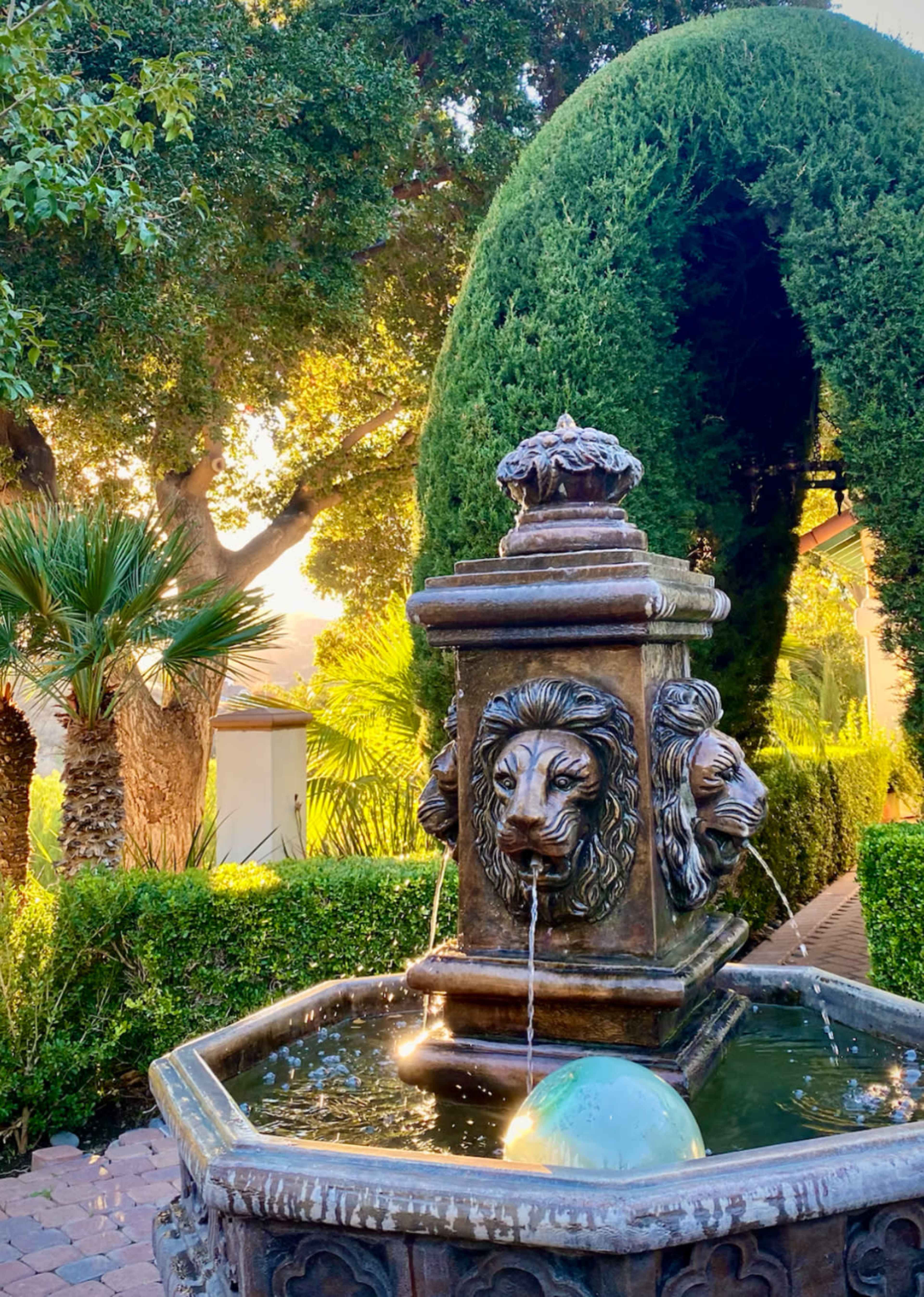 A stone fountain featuring lionhead sculptures spouts water into a basin surrounded by lush greenery and palm trees.