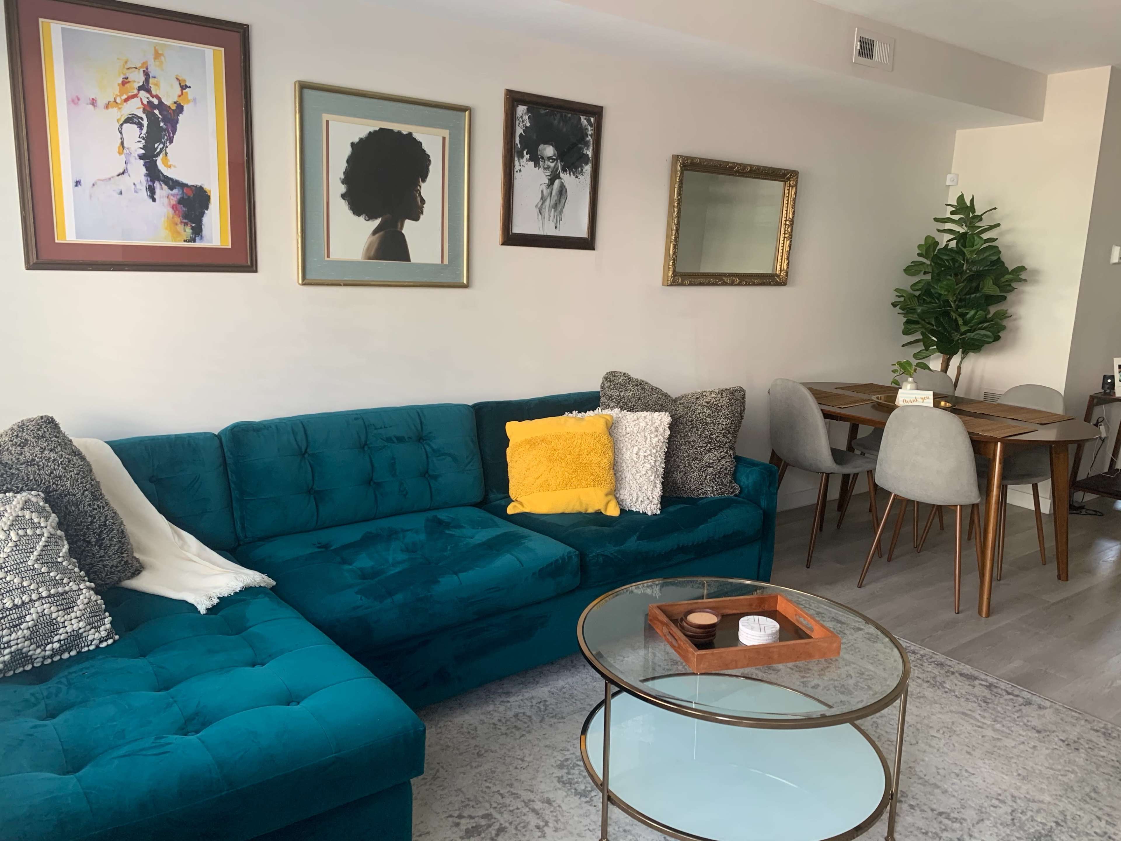 The image shows a modern living room with a teal sectional sofa, a glass coffee table, framed artwork on the walls, and a dining area with a wooden table and gray chairs.