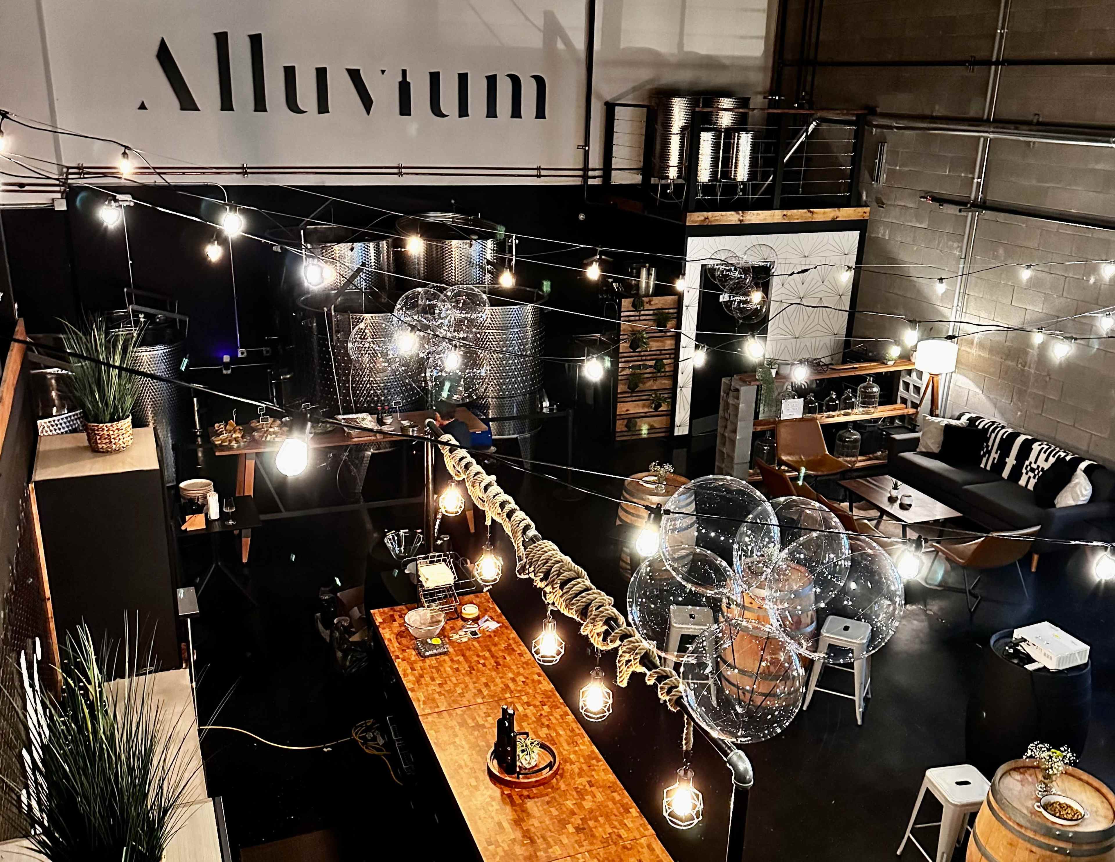 A spacious industrial-style room features a long wooden table adorned with plants and lights, surrounded by modern seating and large storage tanks in the background.