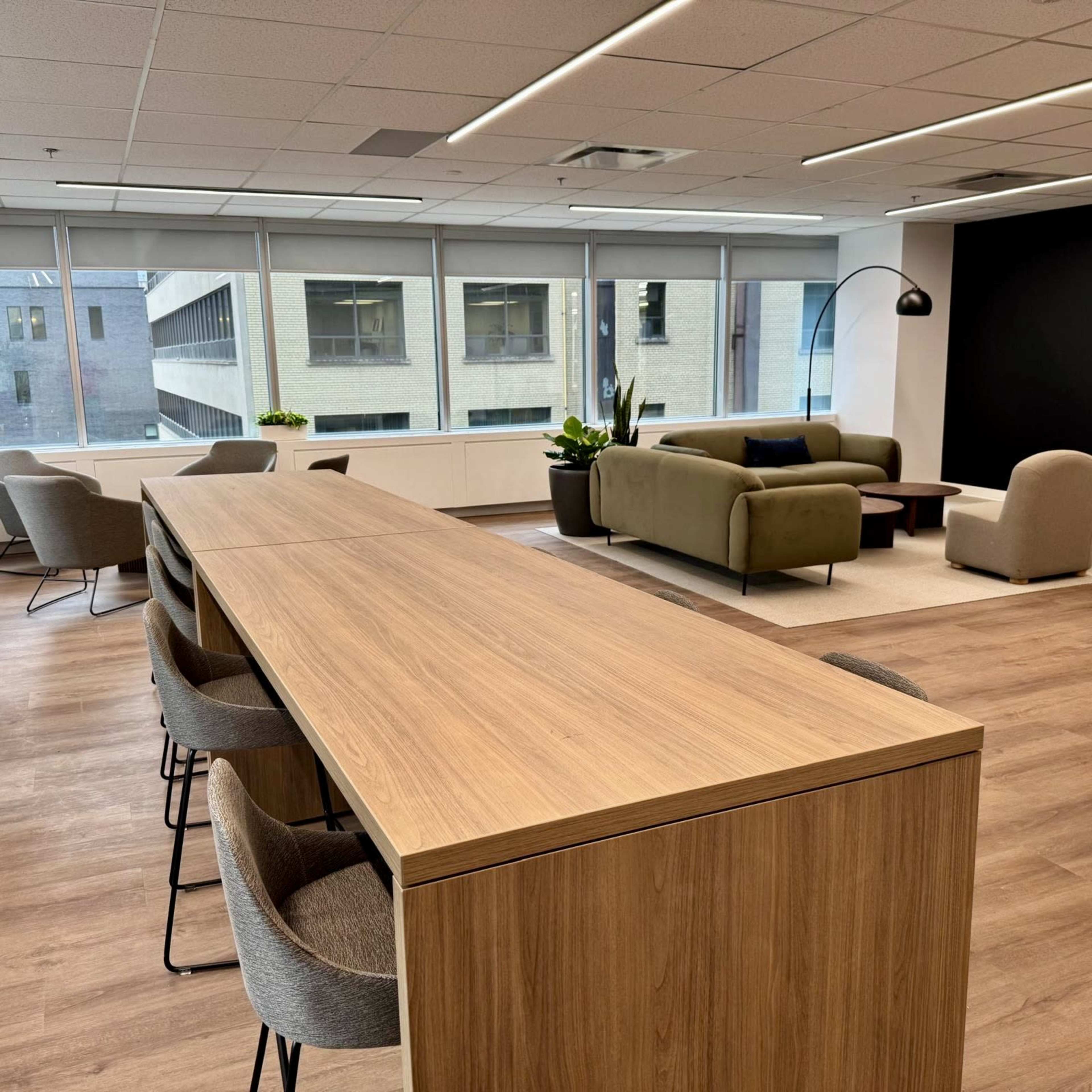The image shows a modern office space featuring a long wooden table with upholstered chairs, large windows allowing natural light, and comfortable seating areas with plants.