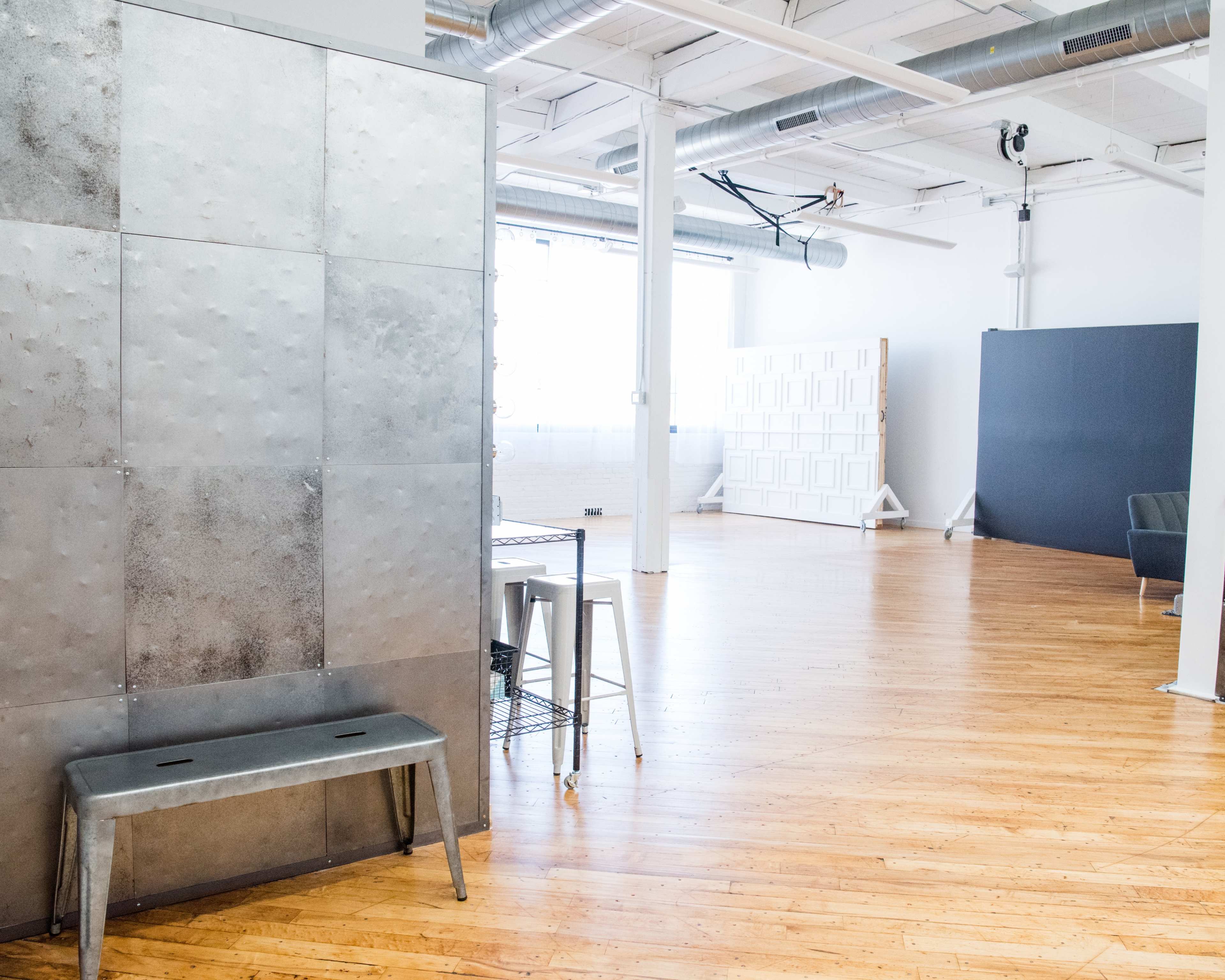 An industrial-style room features wooden floors and large windows, with a metallic partition, a bench, and minimal furniture in the foreground.