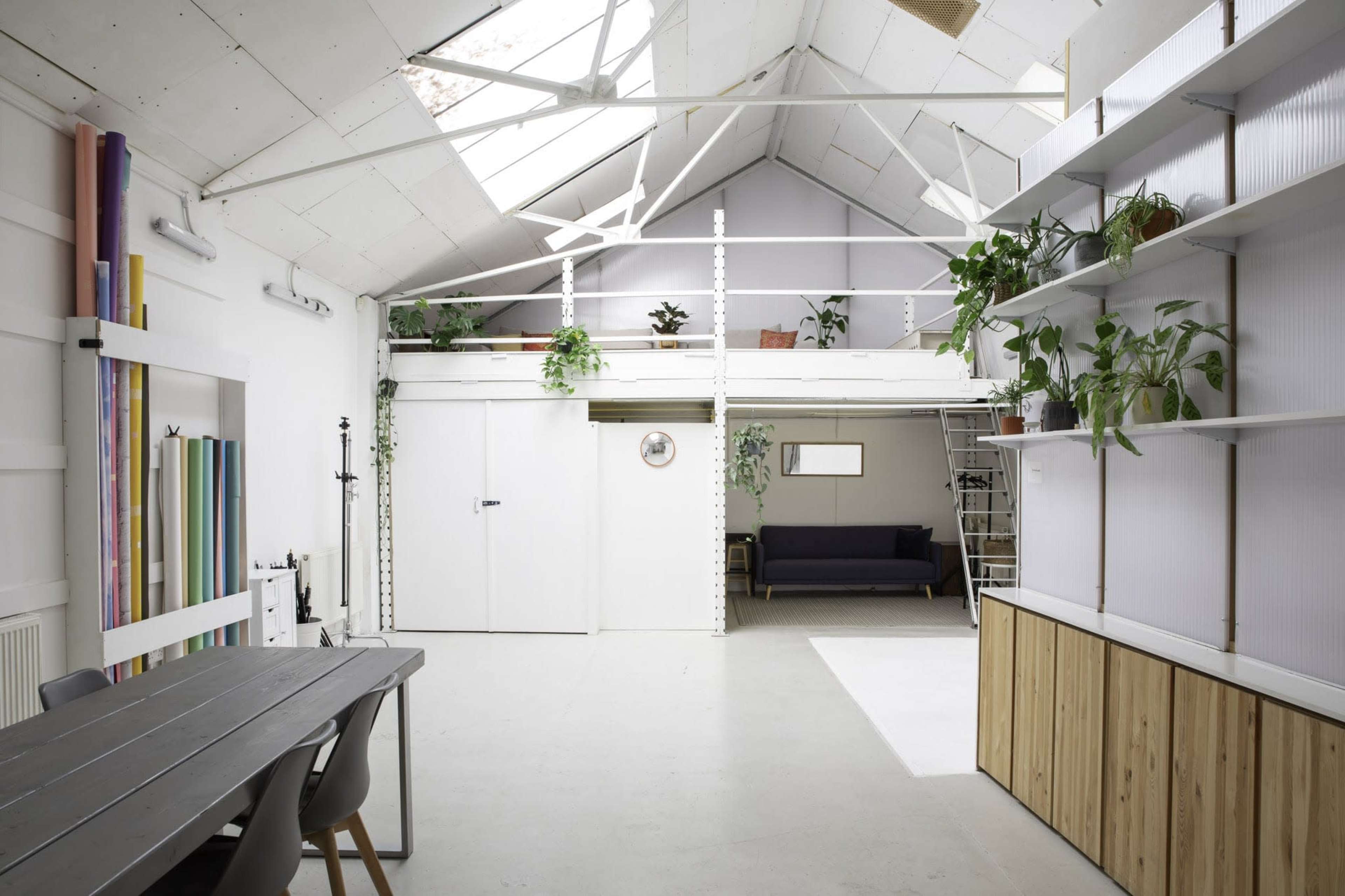 The image shows a modern, open interior of a loft-style space with a high ceiling, plants on shelves, and a seating area in the background.