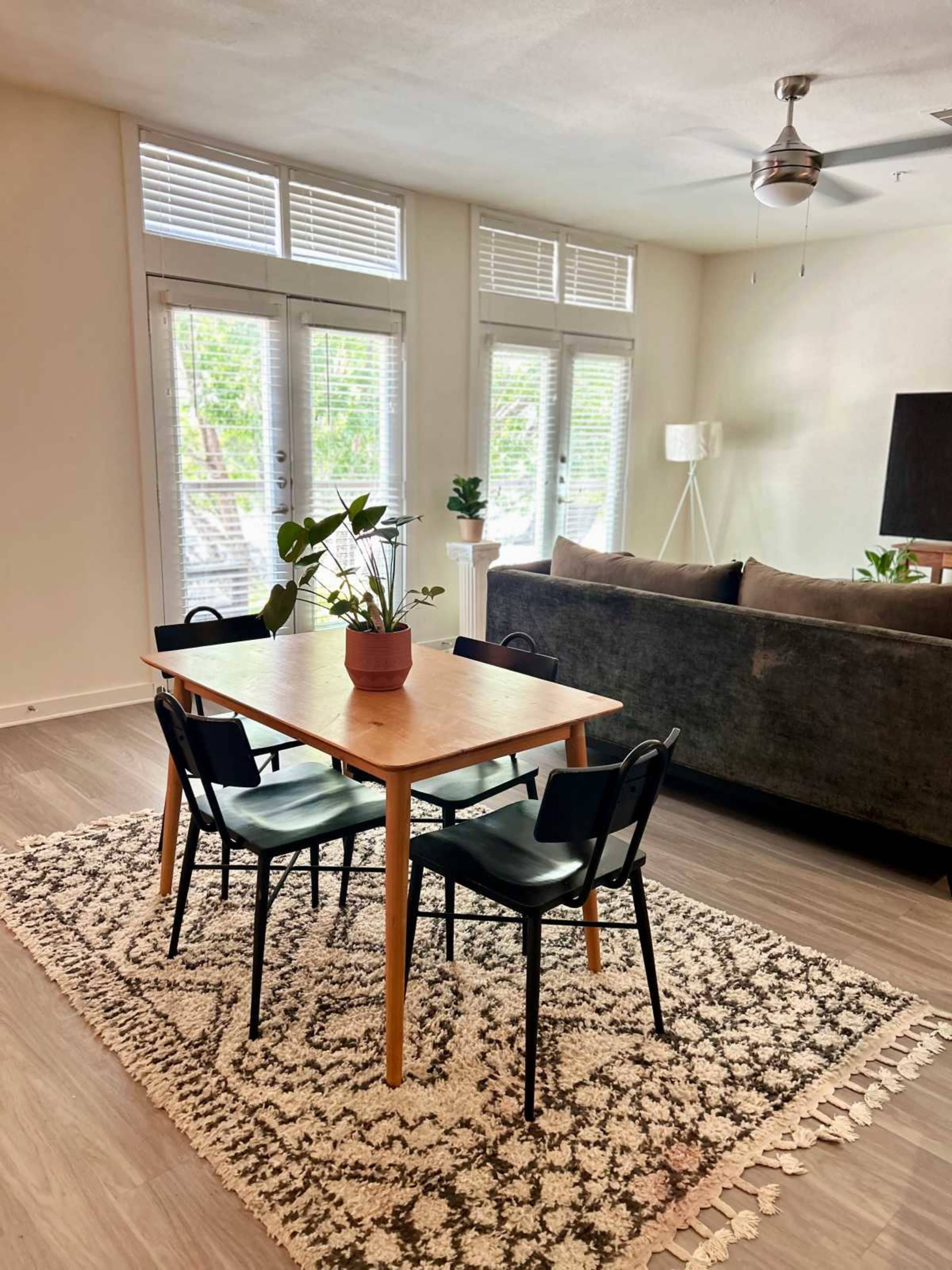 The image shows a dining area with a wooden table surrounded by black chairs, a potted plant on the table, and a gray sofa in the background, with large windows letting in light.