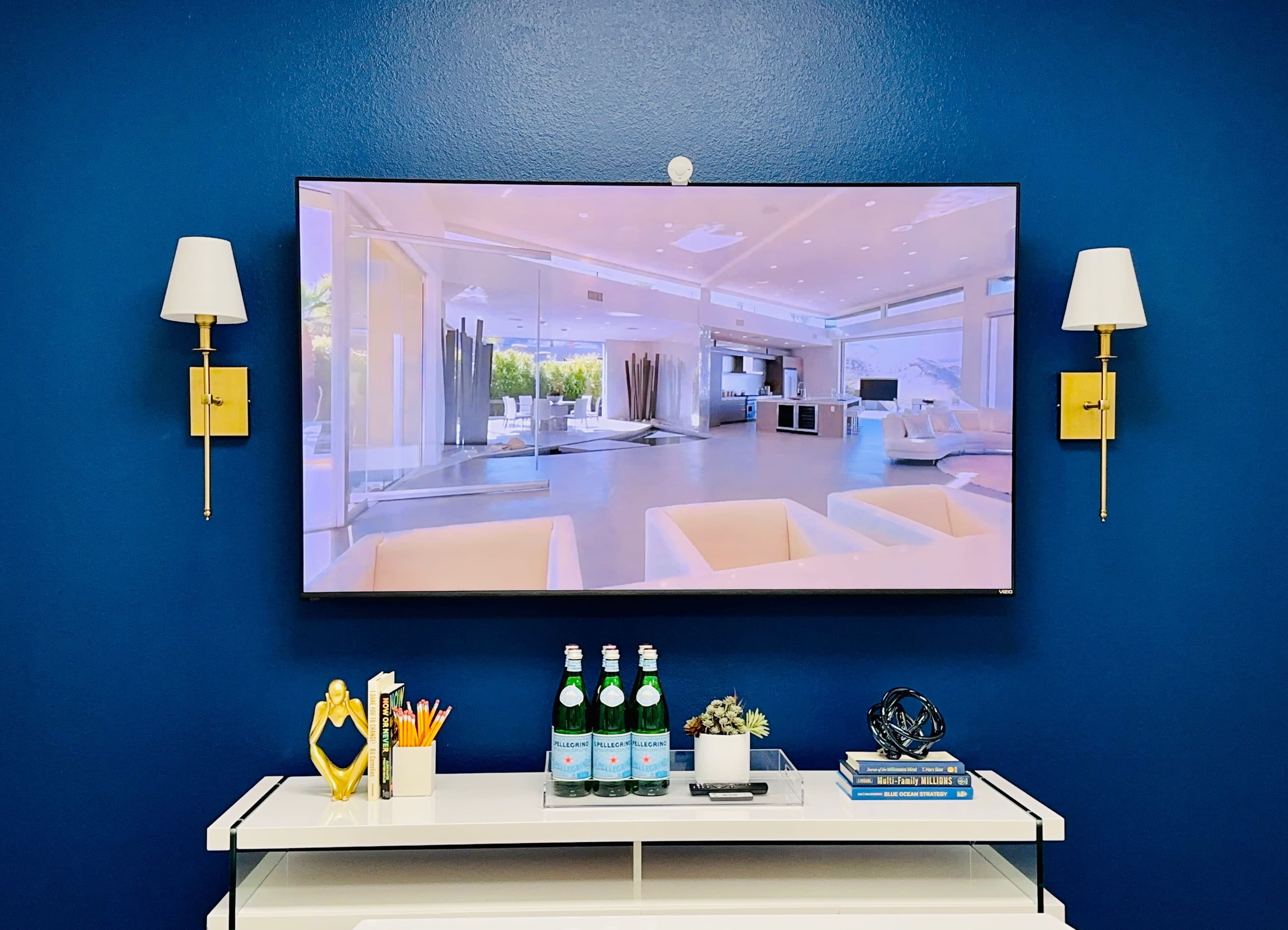 A large flat-screen TV displays a modern interior scene on a blue wall, above a white console table holding decorative items and drinks.