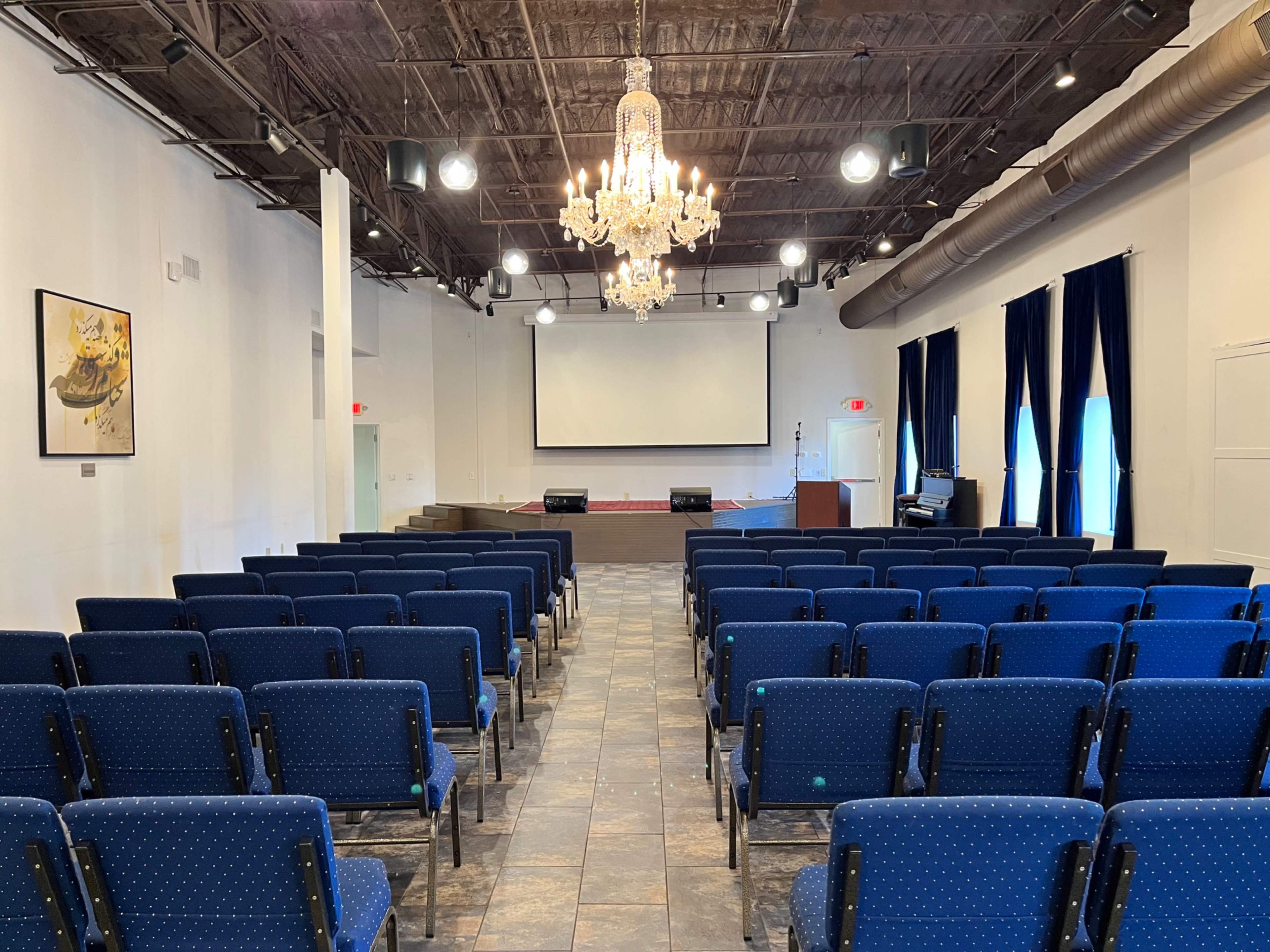 The image depicts a spacious auditorium with rows of blue chairs facing a large screen and a stage at the front, illuminated by a chandelier overhead.