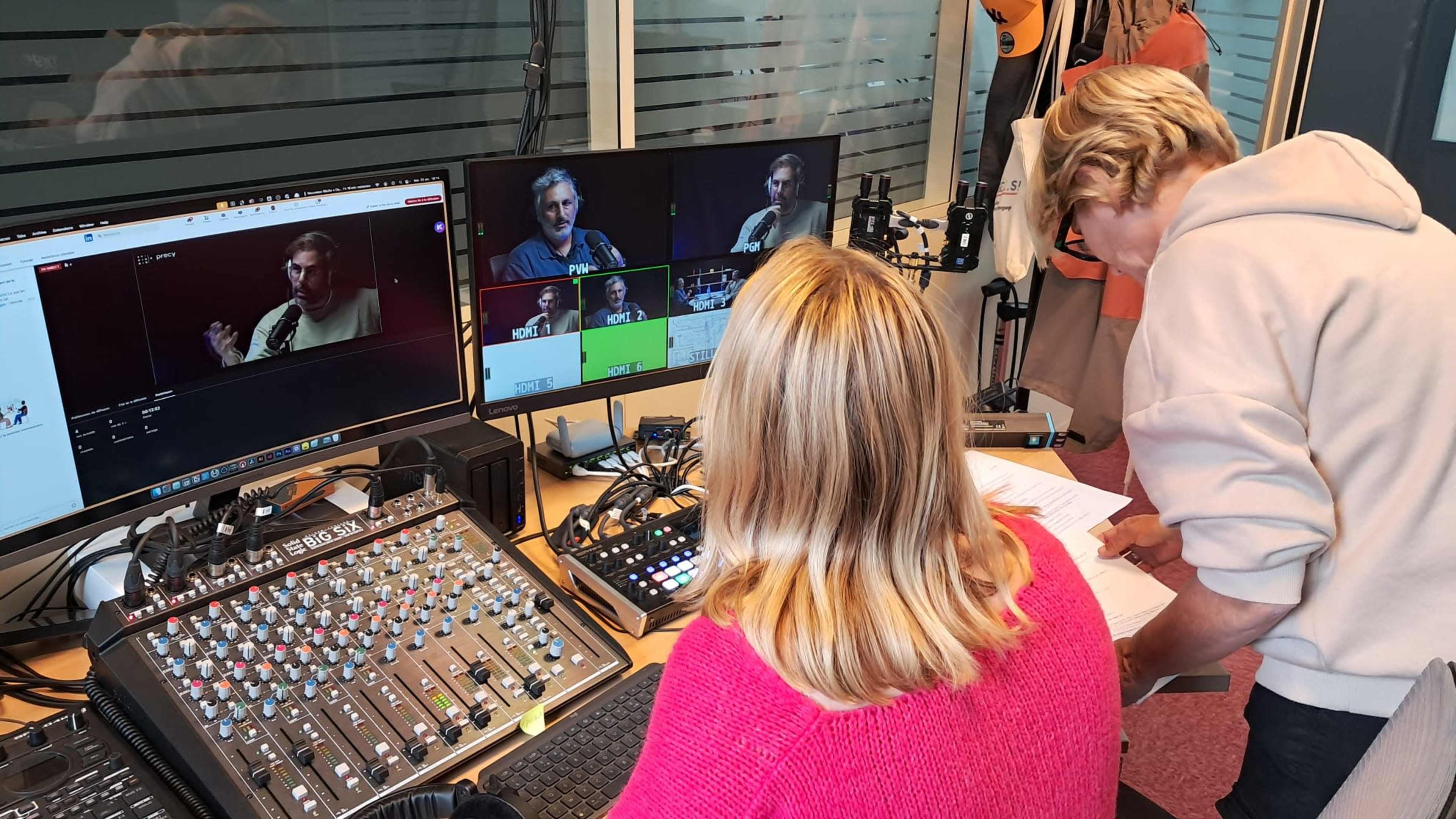 A person adjusts equipment at a video production station while another individual monitors multiple screens displaying live video feeds.