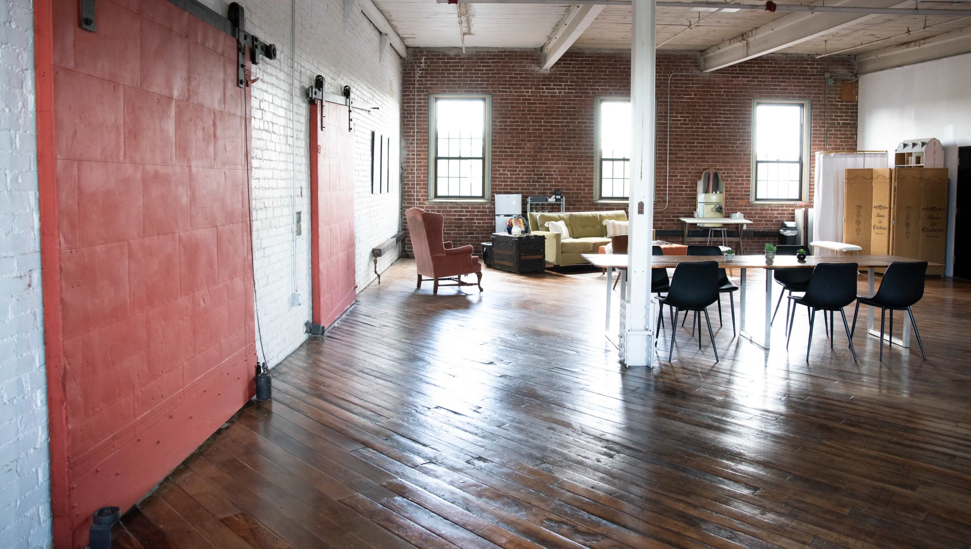 A spacious, empty room with wooden floors, large windows, and a dining area featuring a table and chairs, alongside a section with seating and cardboard boxes.