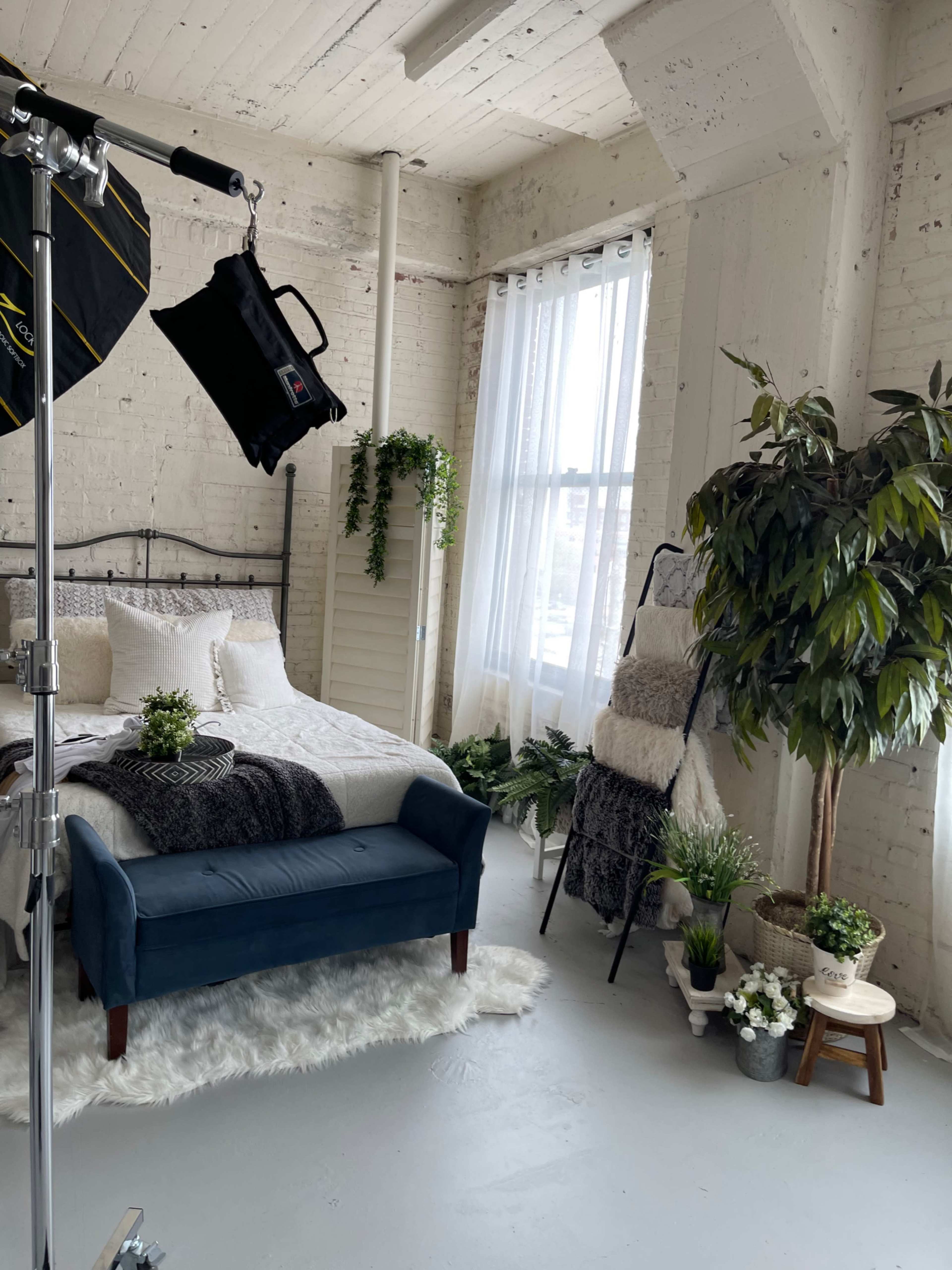 A well-lit bedroom setup with a bed, a blue sofa, and decorative plants, featuring a white-washed brick wall and large windows.