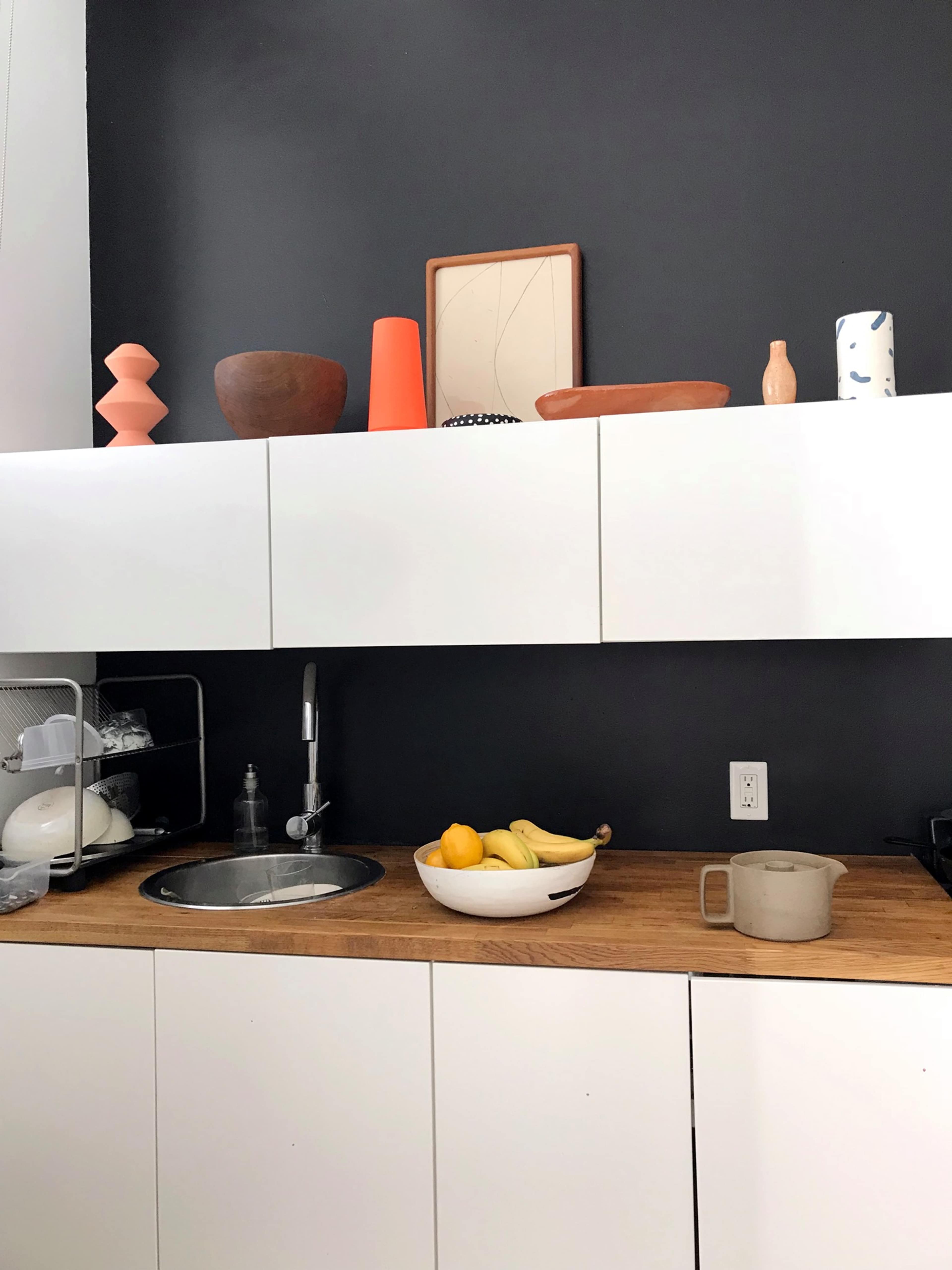 A modern kitchen features a wooden countertop with a basin, a fruit bowl, and various decorative items arranged on a shelf above white cabinets.
