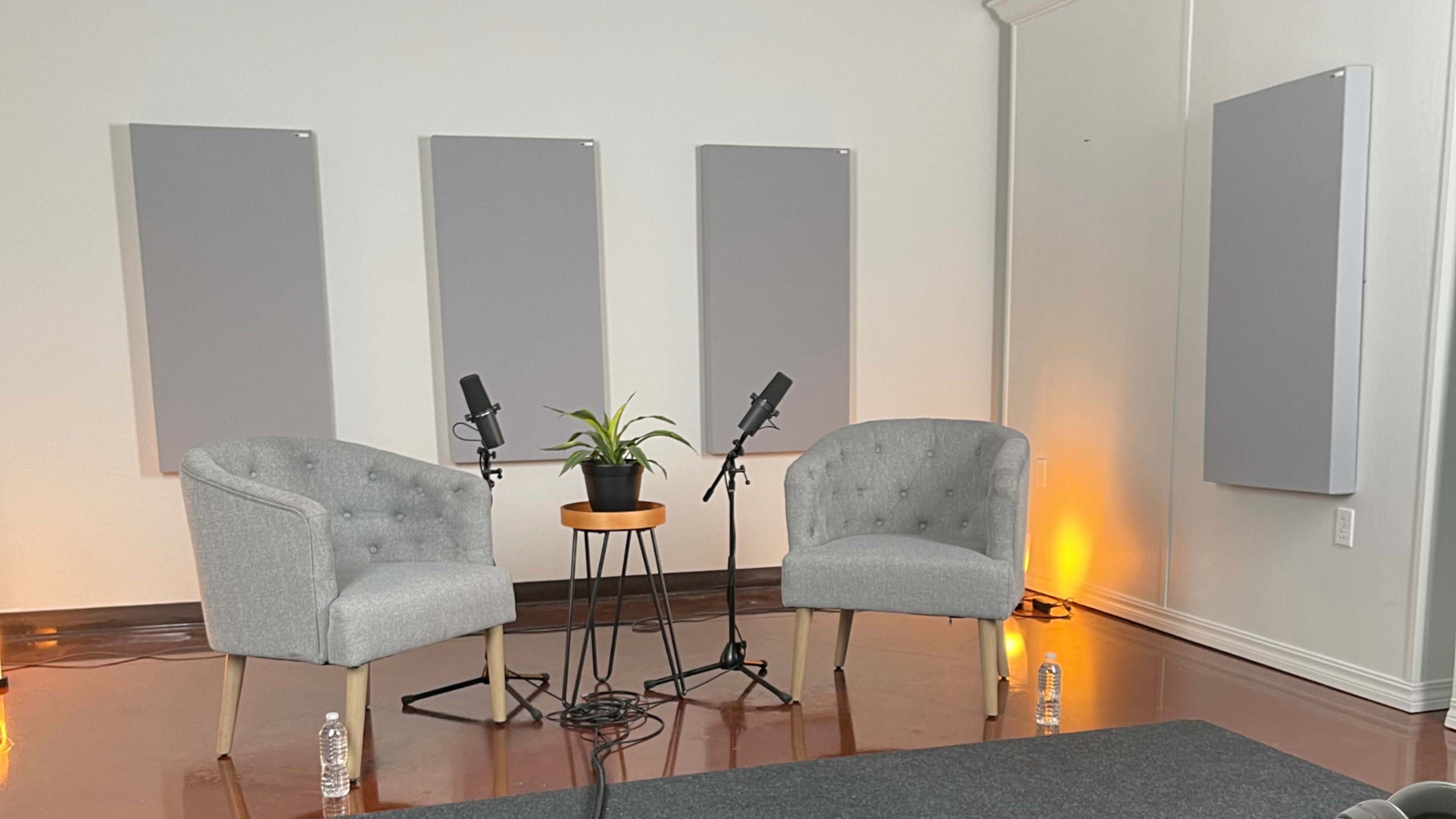 Two gray upholstered chairs facing each other on a carpeted floor, with a small table featuring a potted plant and microphones positioned between them, against a backdrop of four sound-absorbing panels on the wall.
