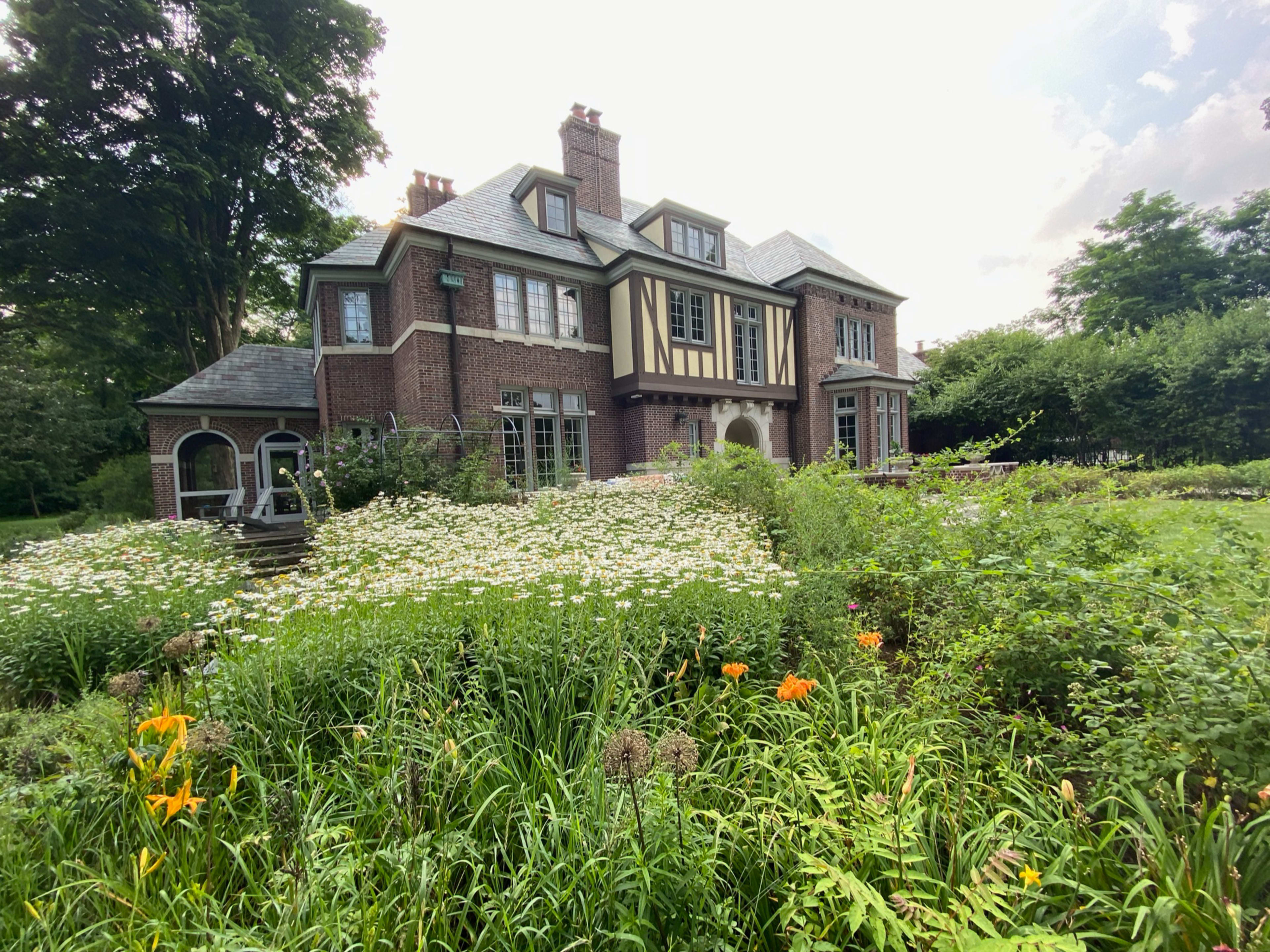 A large, brick house with a gabled roof is surrounded by a garden filled with various flowers and greenery.