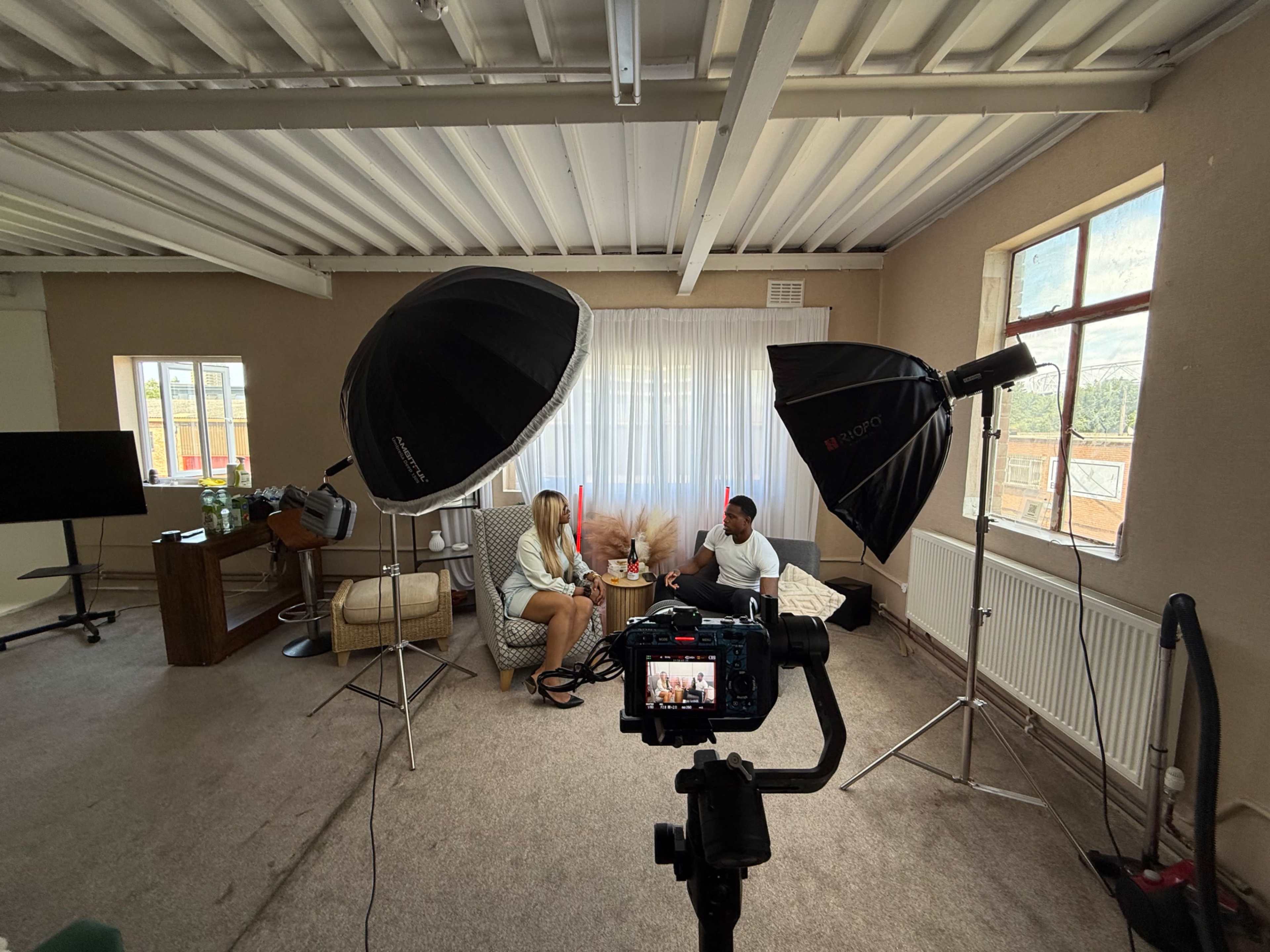 The image shows two people seated in a well-lit room, surrounded by photography and lighting equipment.