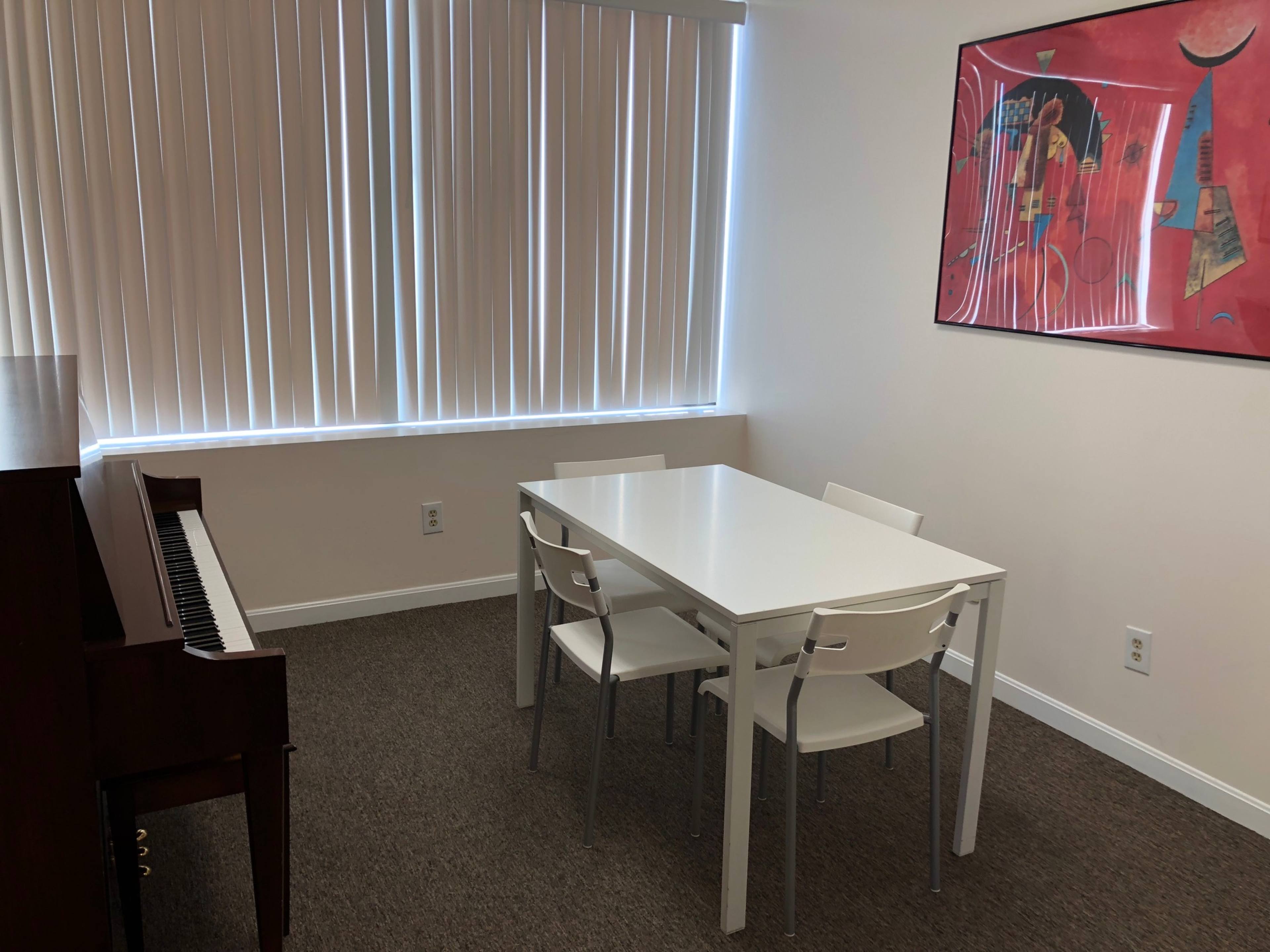 A white table with four chairs is set against a wall with a window and a colorful abstract painting, next to a brown piano.