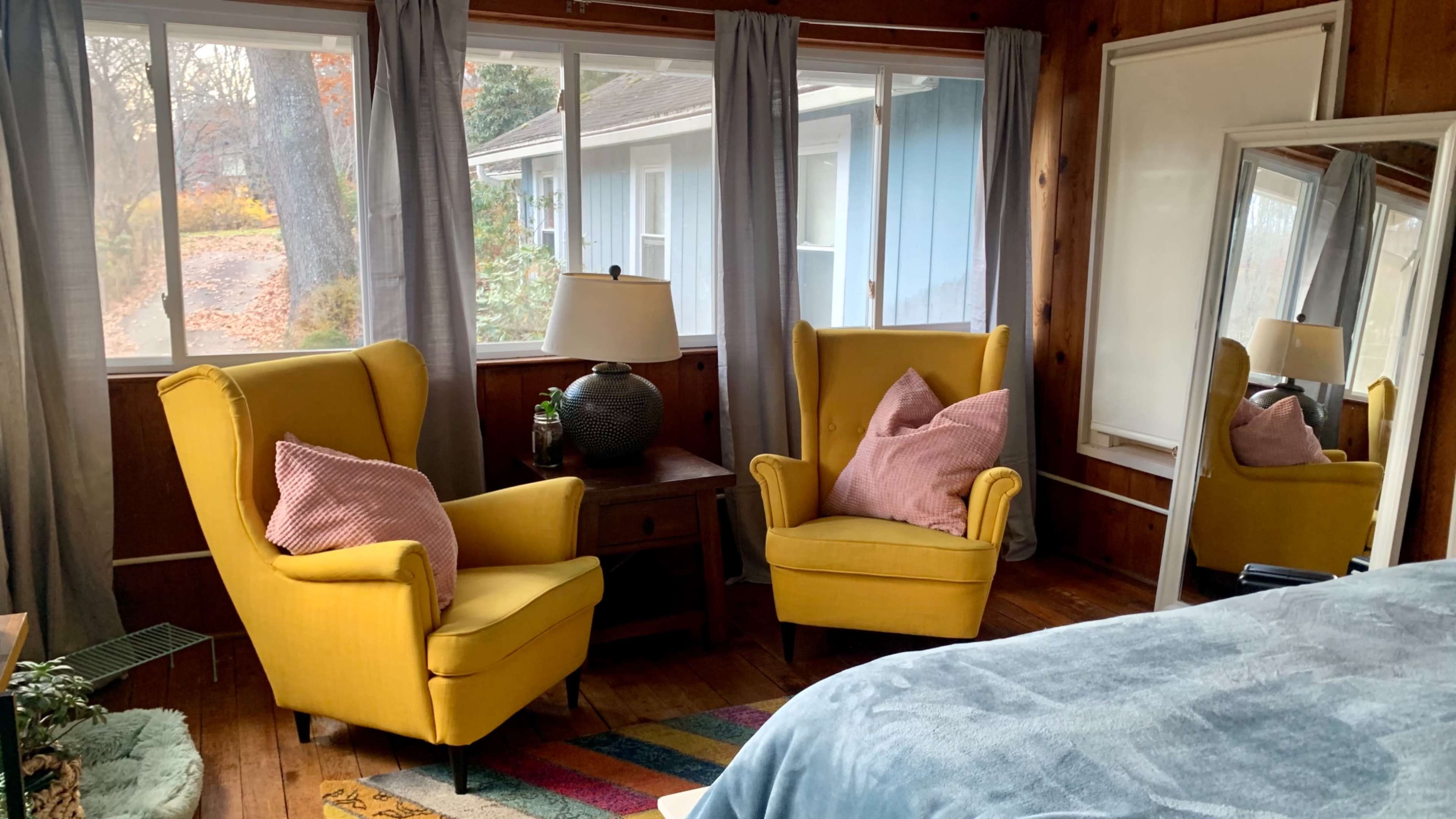 The image shows a cozy room with two yellow armchairs, a wooden nightstand with a lamp, and a large mirror, all surrounded by large windows.