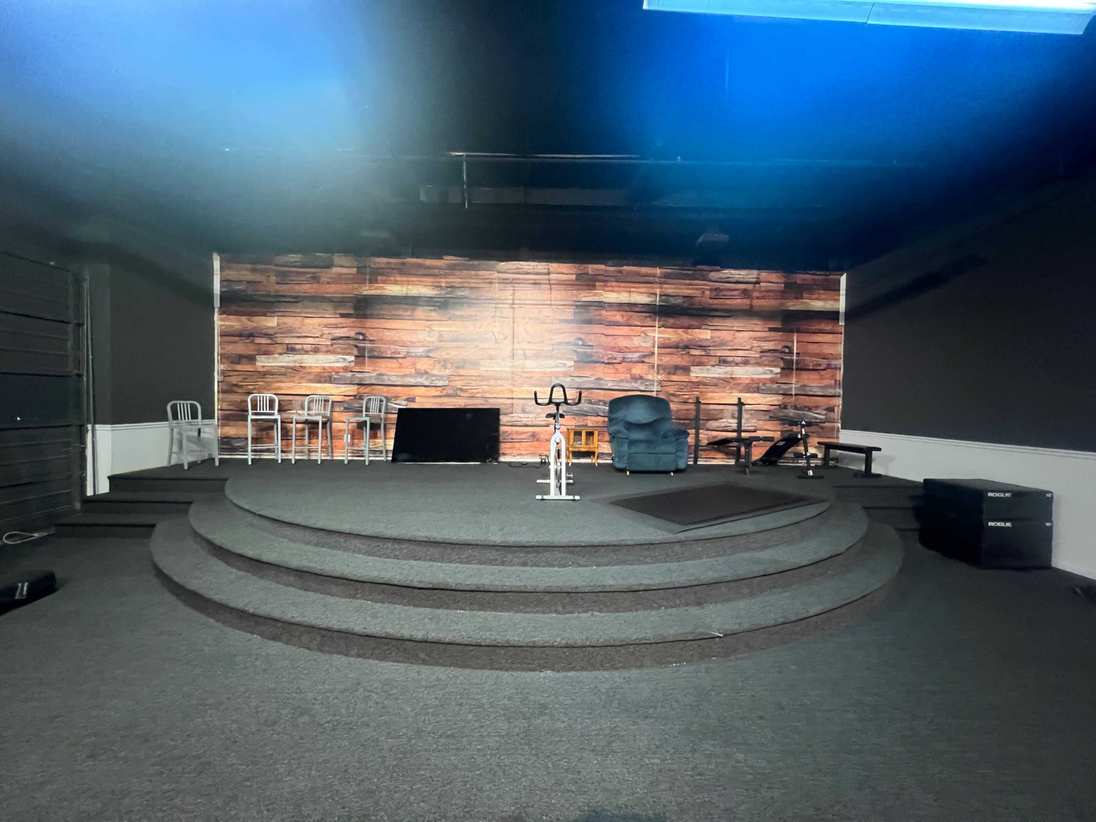 The image shows a stage with a wooden backdrop, circular steps, a stationary bike, a set of chairs, and a few fitness equipment pieces in a dimly lit room.