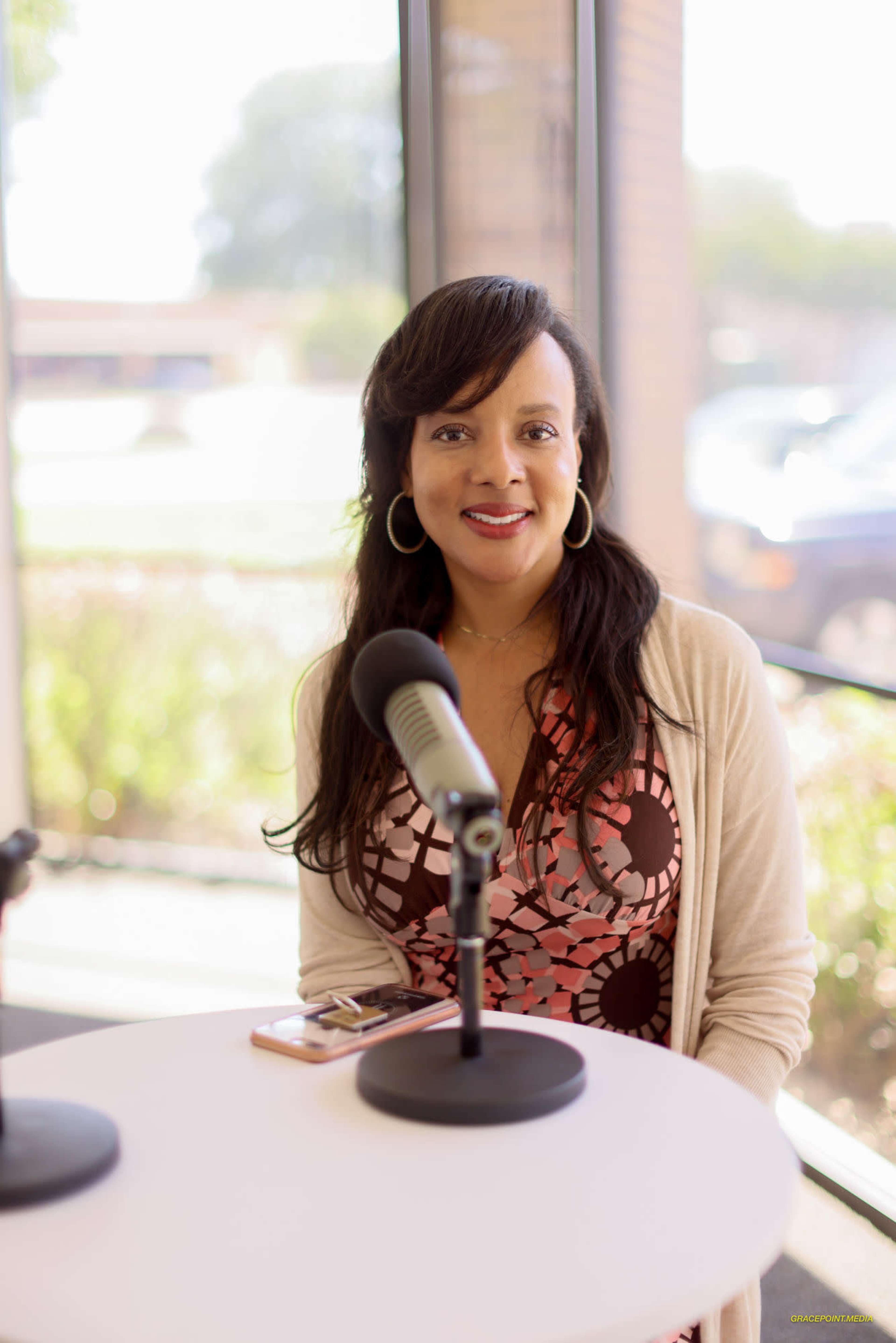 A woman with long brown hair sits at a table with a microphone in front of her, smiling and looking towards the camera.