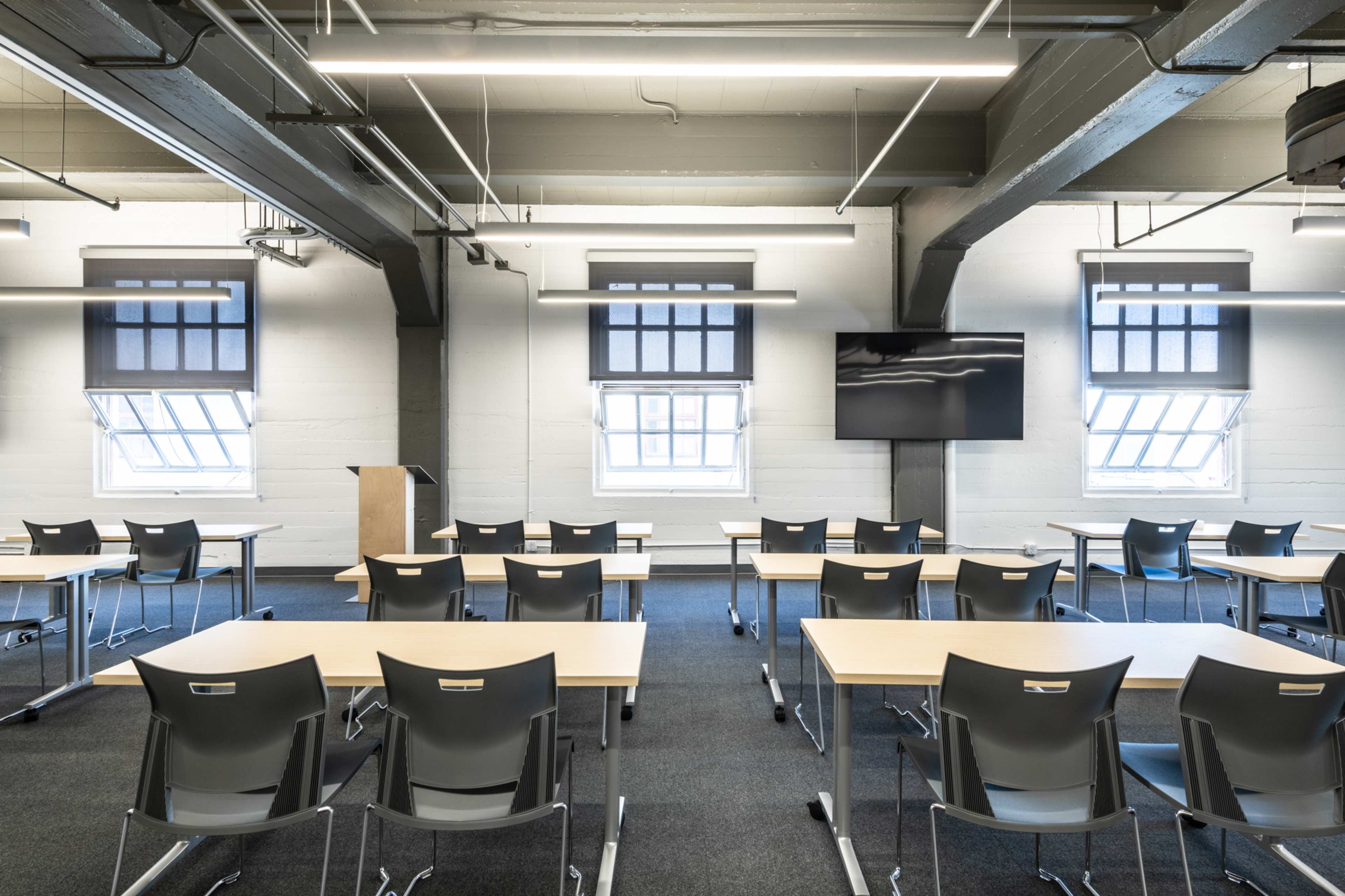 A modern classroom with rows of tables and chairs, large windows, and a wall-mounted screen at the front.