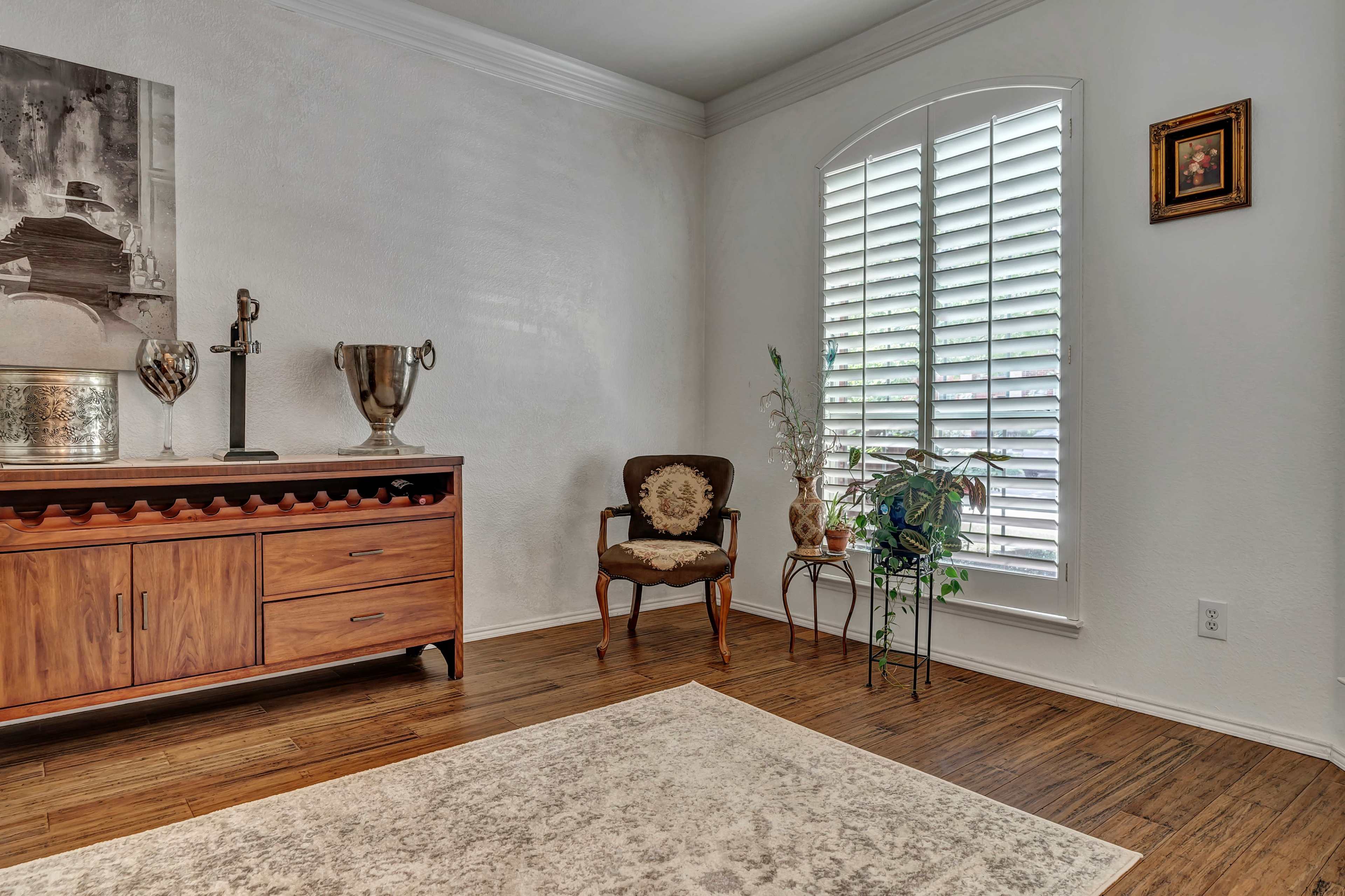 A wooden sideboard is positioned against a wall in a room with a window featuring white shutters, a chair in the corner, and a small potted plant.