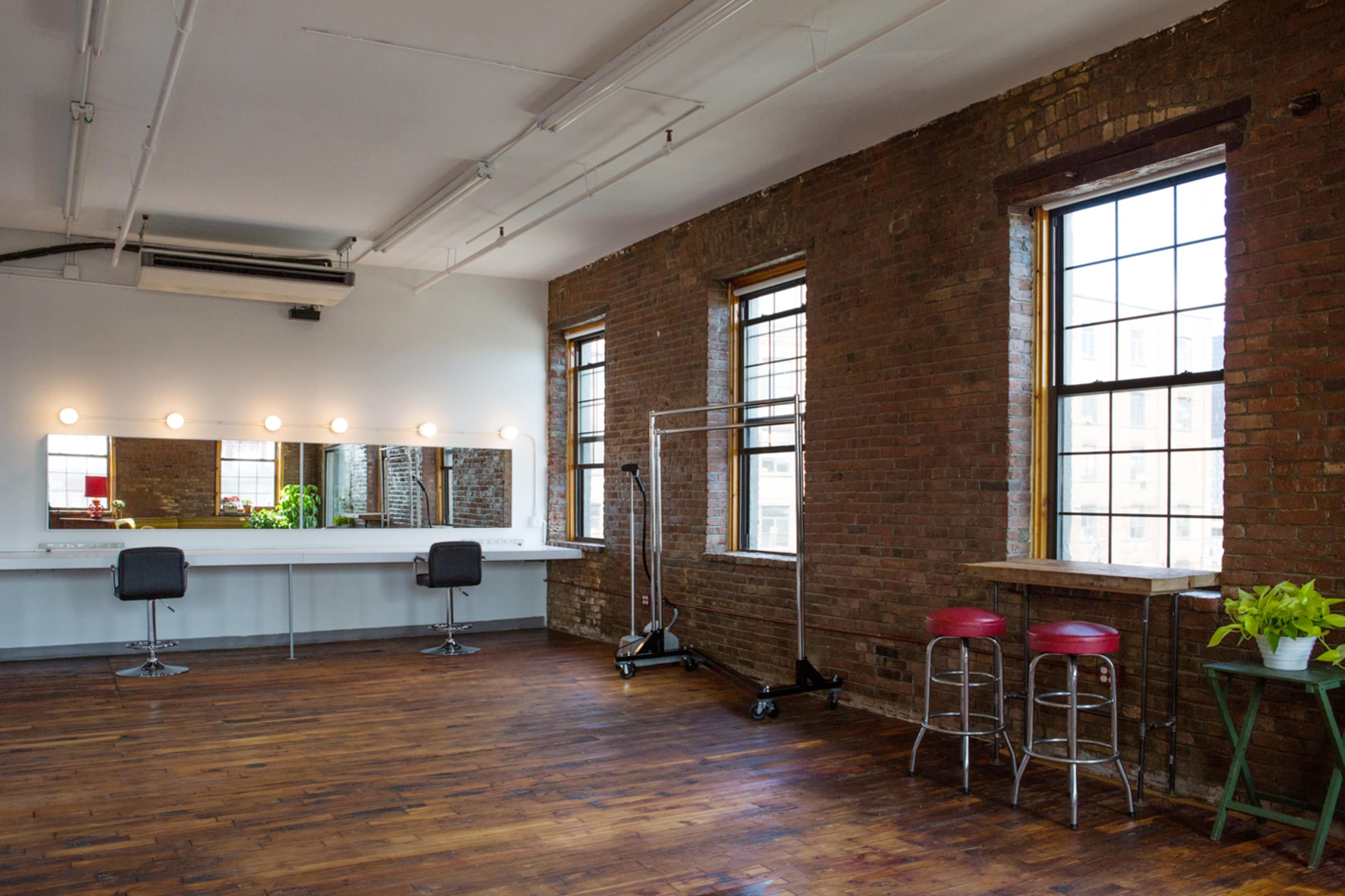A bright, spacious room with exposed brick walls, a large mirror with lights, and a wooden floor, furnished with a seating area and a clothing rack.