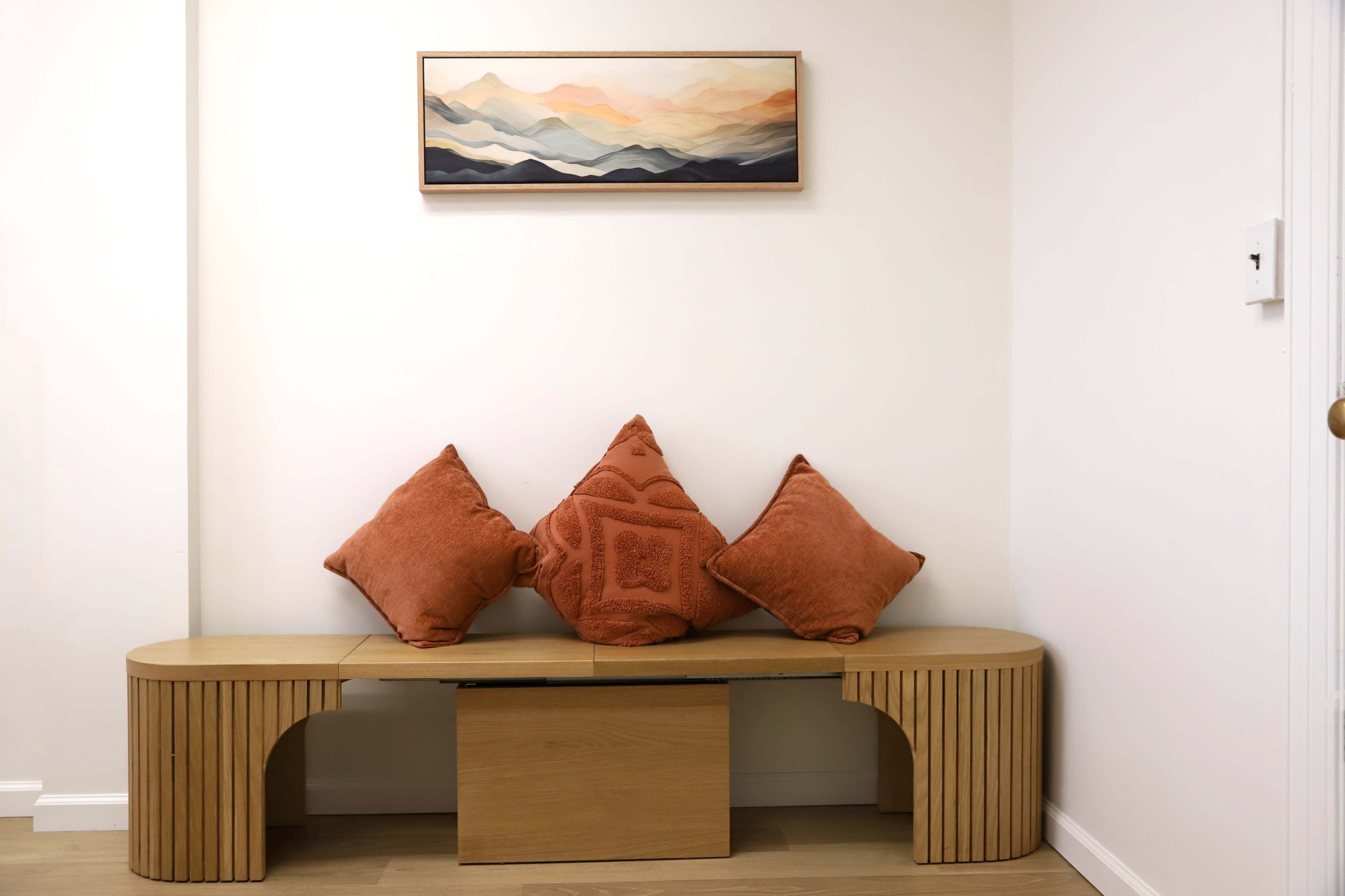 The image shows a minimalist interior scene featuring a wooden bench with three orange pillows and a mountain landscape painting on the wall above.