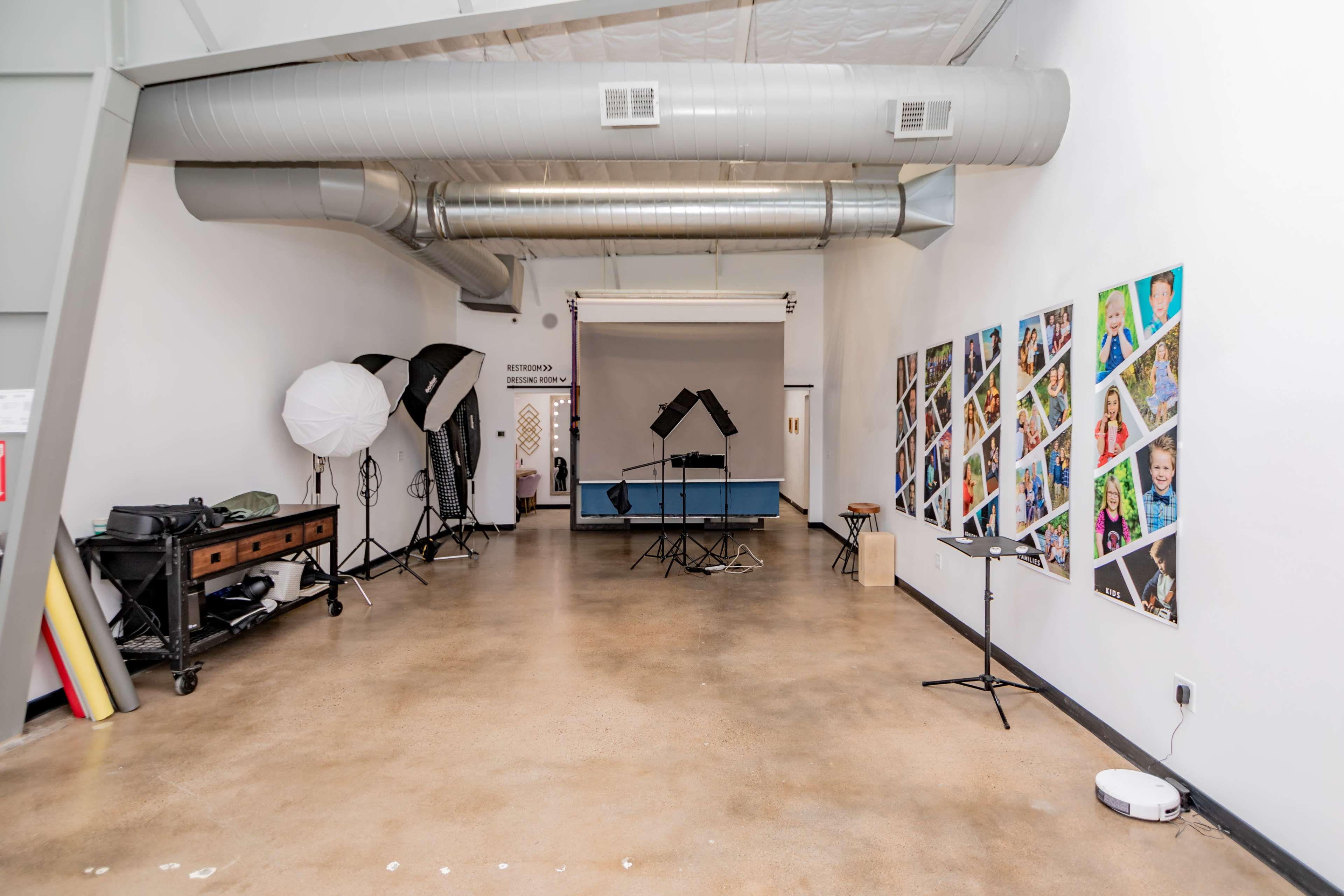 The image shows a photography studio with equipment, including softboxes and backdrops, and a wall displaying various framed photos.