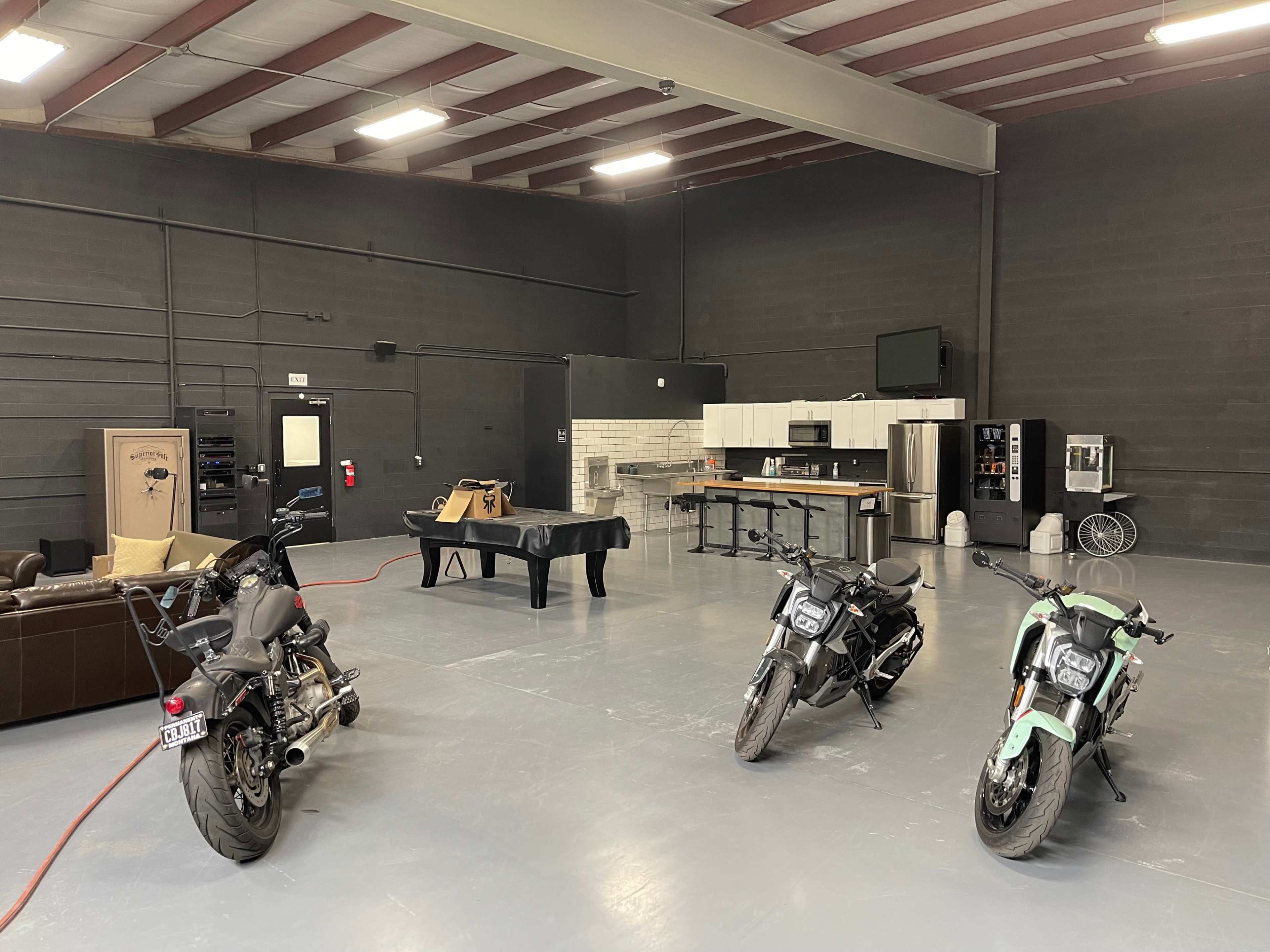 An industrial-style garage with three motorcycles, a couch, a table, and a kitchen area.