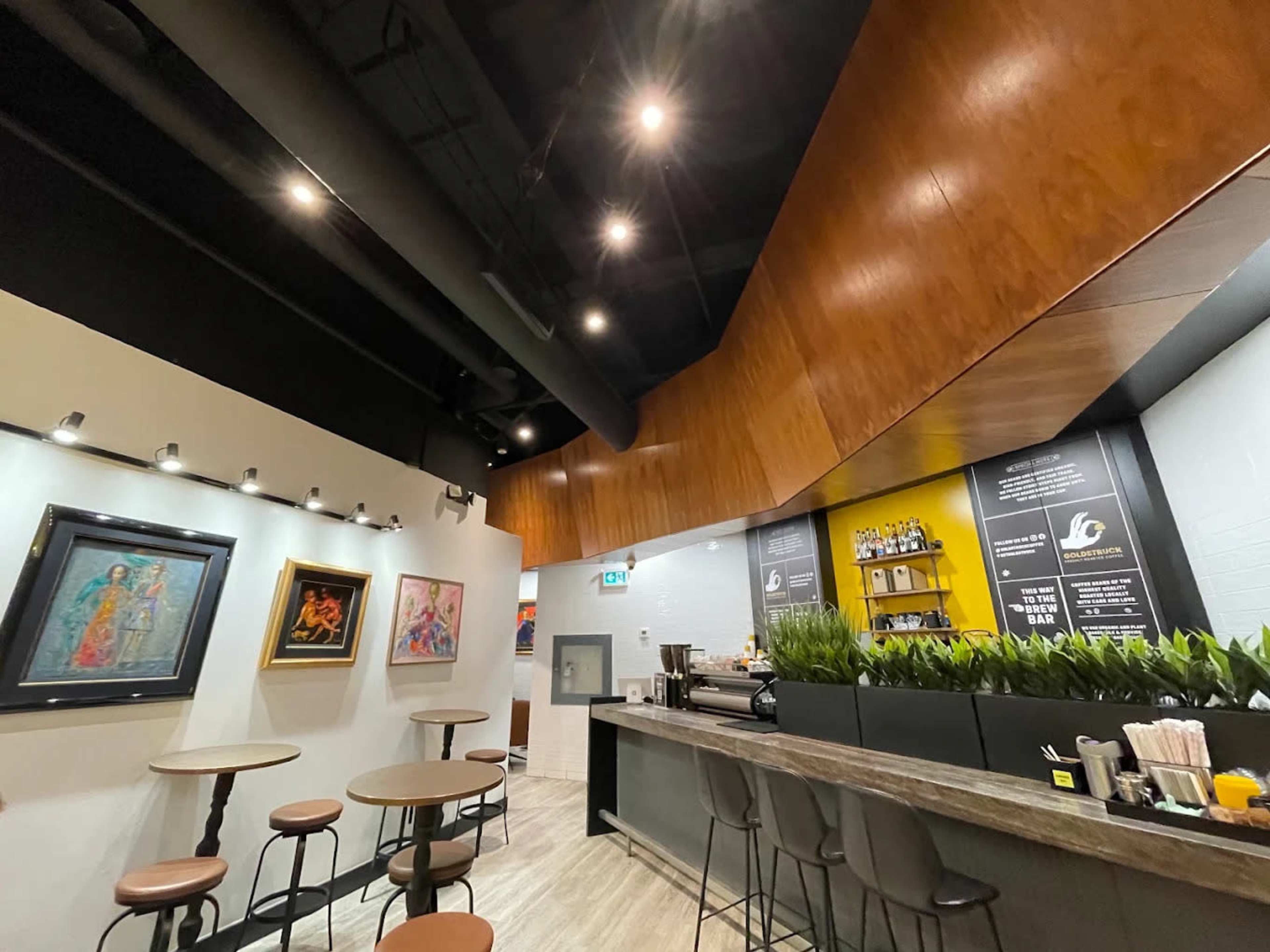 The image shows a modern café interior with a wooden ceiling, potted plants along the bar, and artwork displayed on the walls.