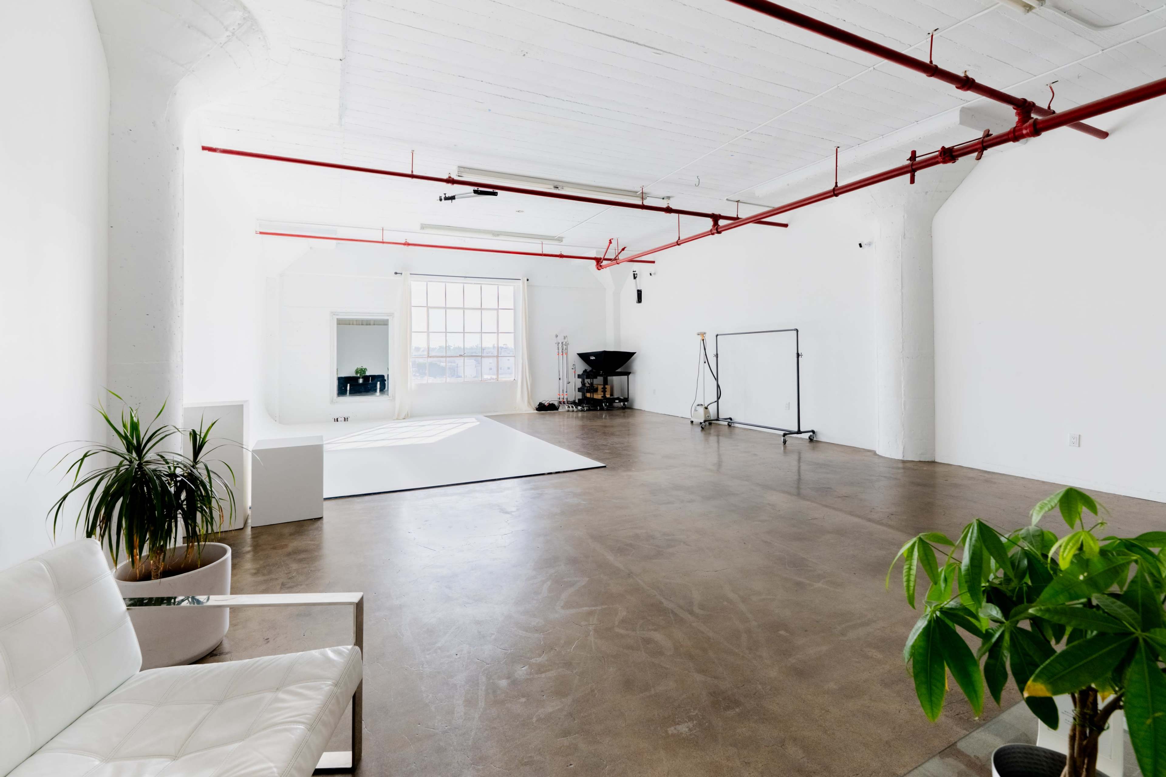The image shows a spacious, bright studio with concrete flooring, white walls, and industrial-style red piping overhead, featuring a seating area, a photography backdrop, and a large window allowing natural light.