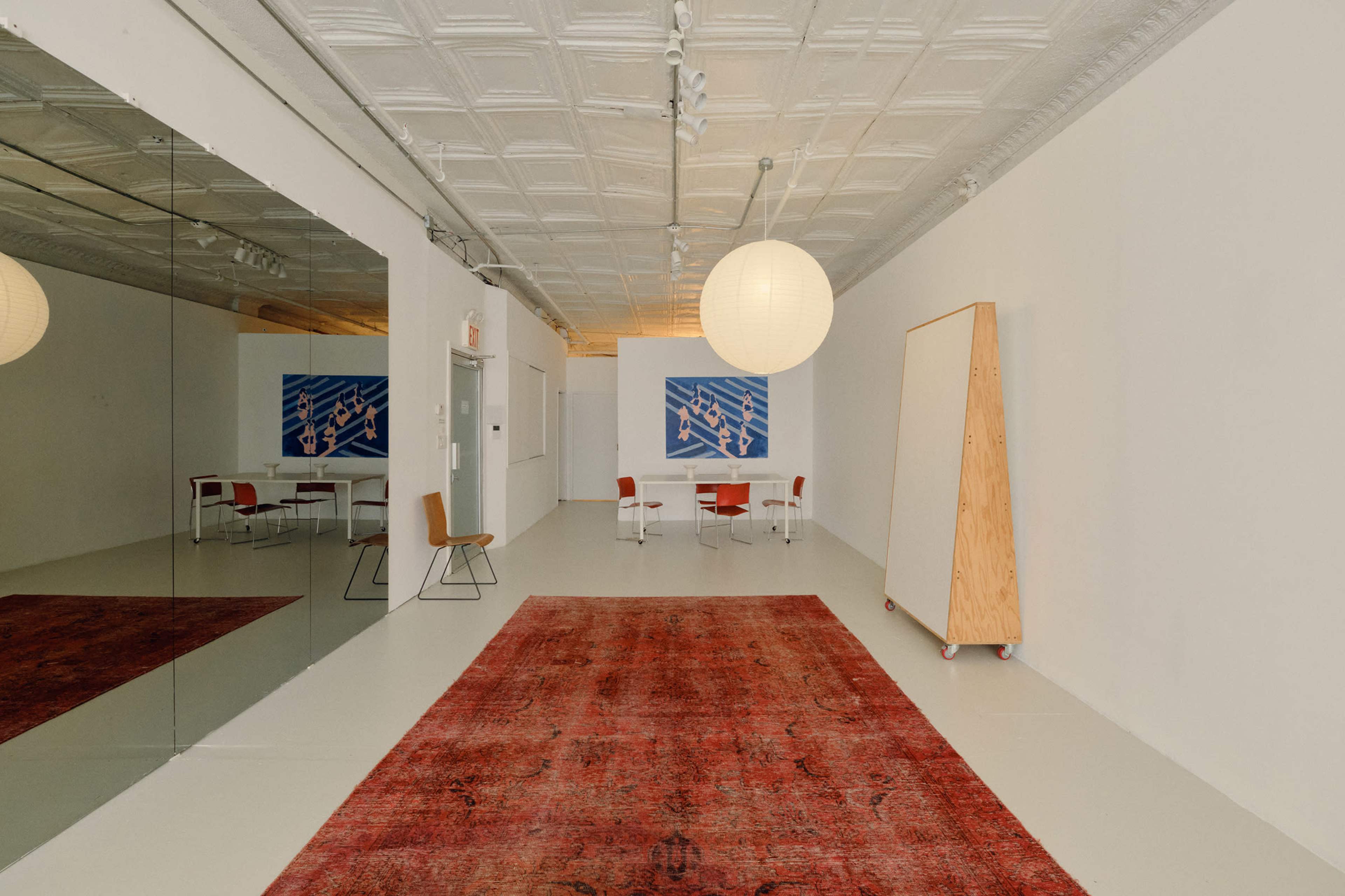 The image depicts a spacious, minimalist room with white walls, a patterned rug, a round pendant light, and a dining area featuring a table and chairs, along with a large mirror on one side.