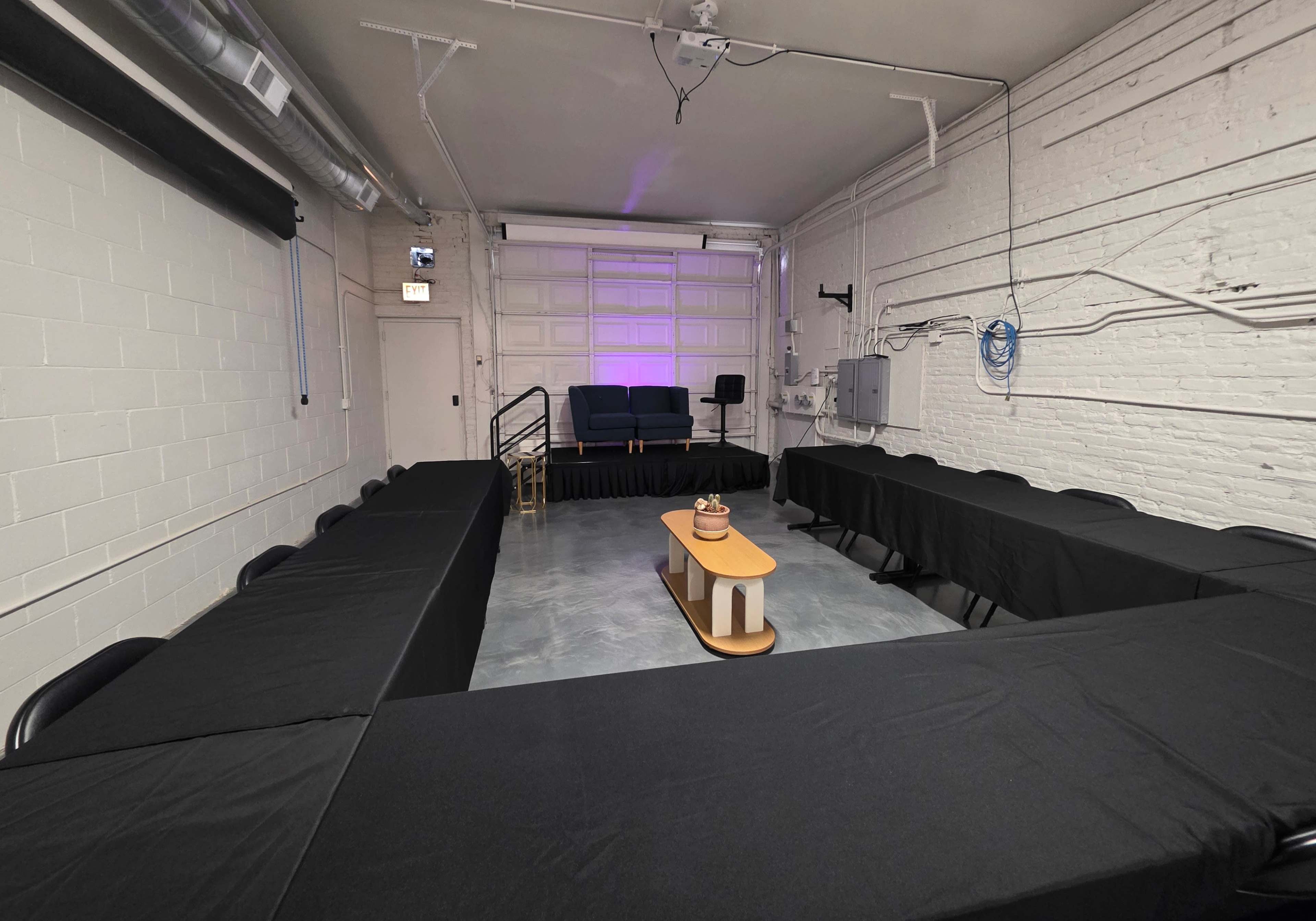 The image shows a meeting room with a rectangular arrangement of tables covered in black cloth, a central coffee table, and a blue couch against a back wall.