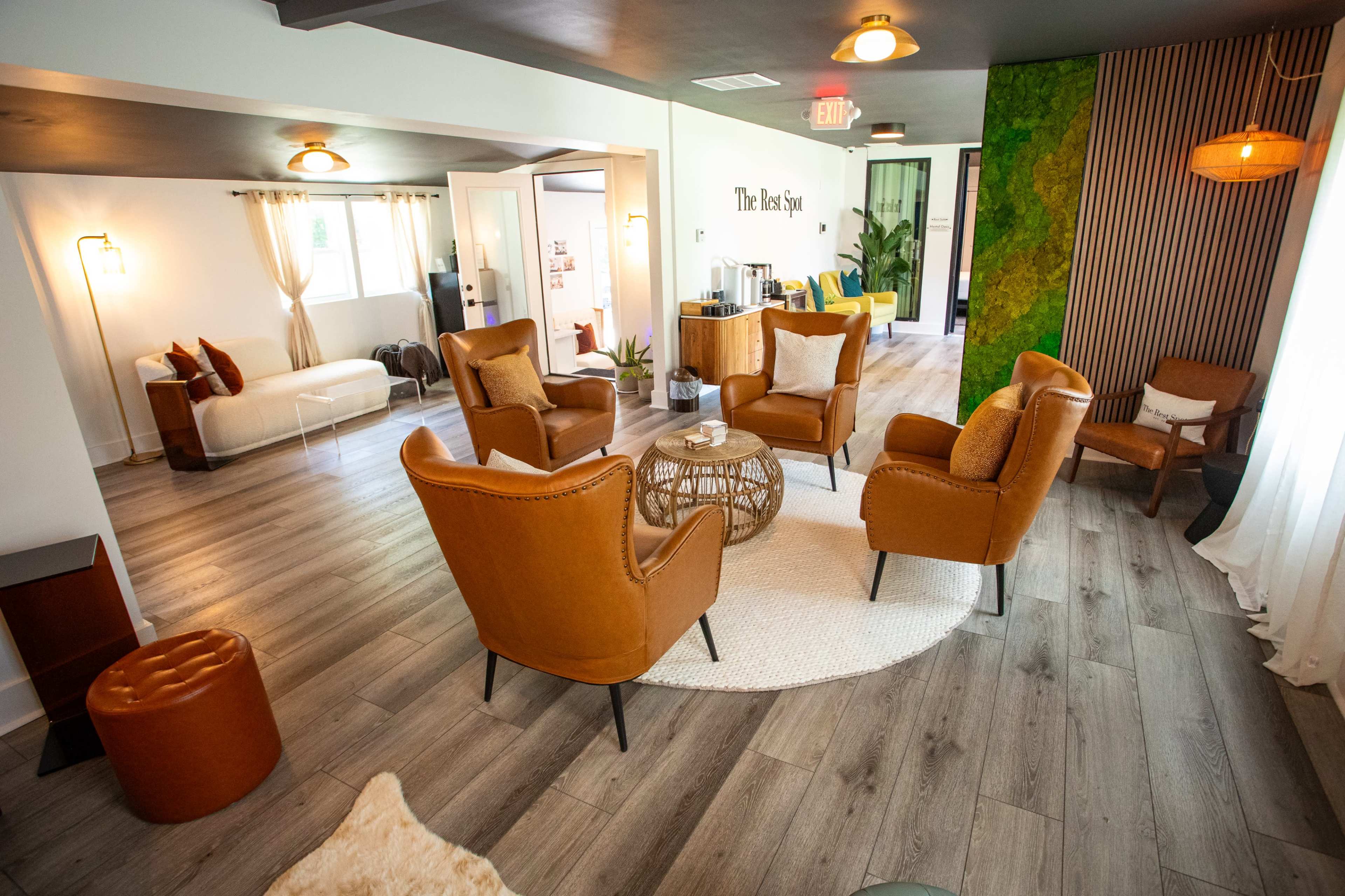 The image shows a modern, well-lit lounge area with four orange armchairs arranged around a small round coffee table on a light-colored rug.