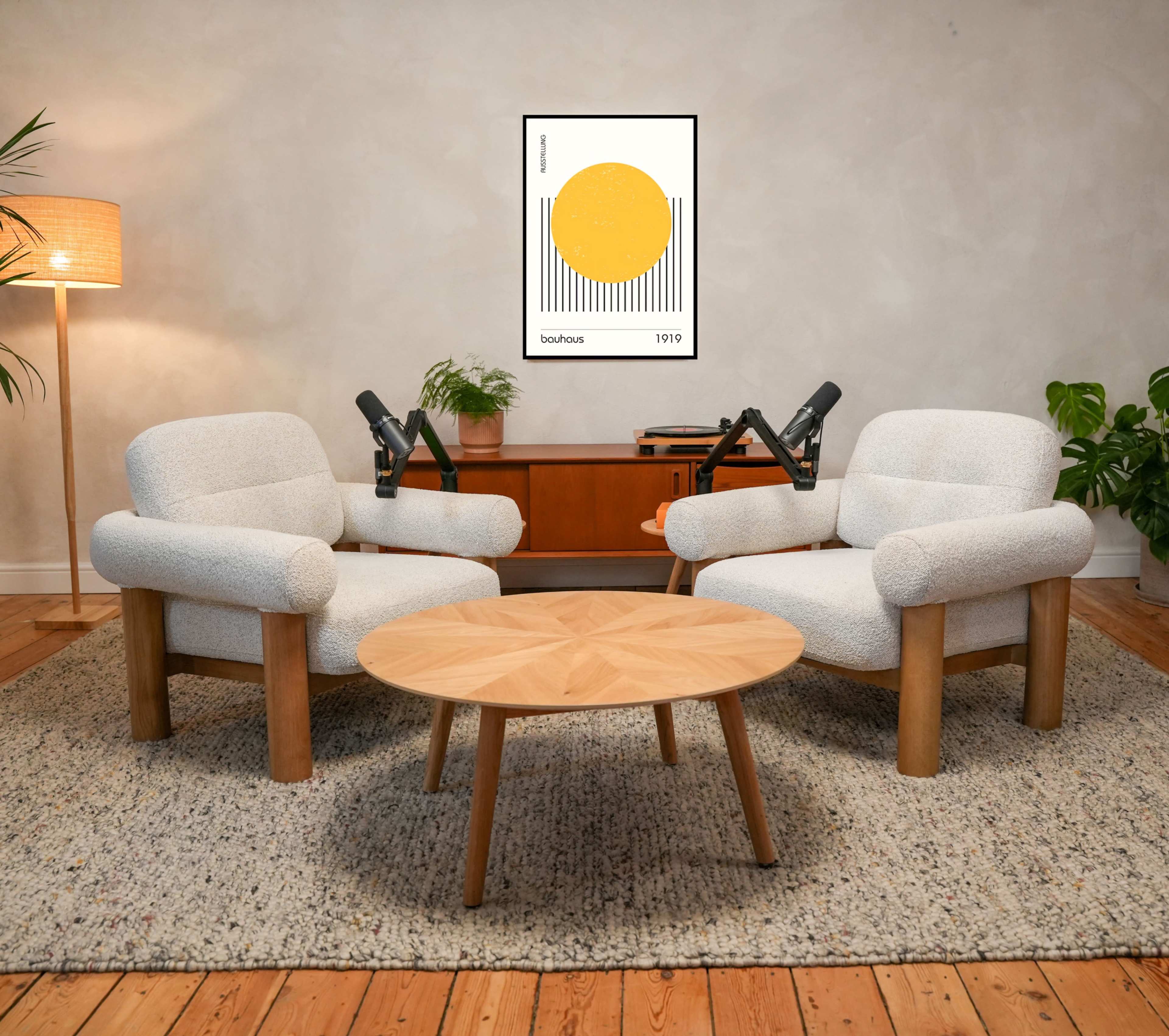 The image shows a cozy interior with two cushioned armchairs facing a wooden coffee table, accompanied by a rug, a floor lamp, and a wall-mounted poster.