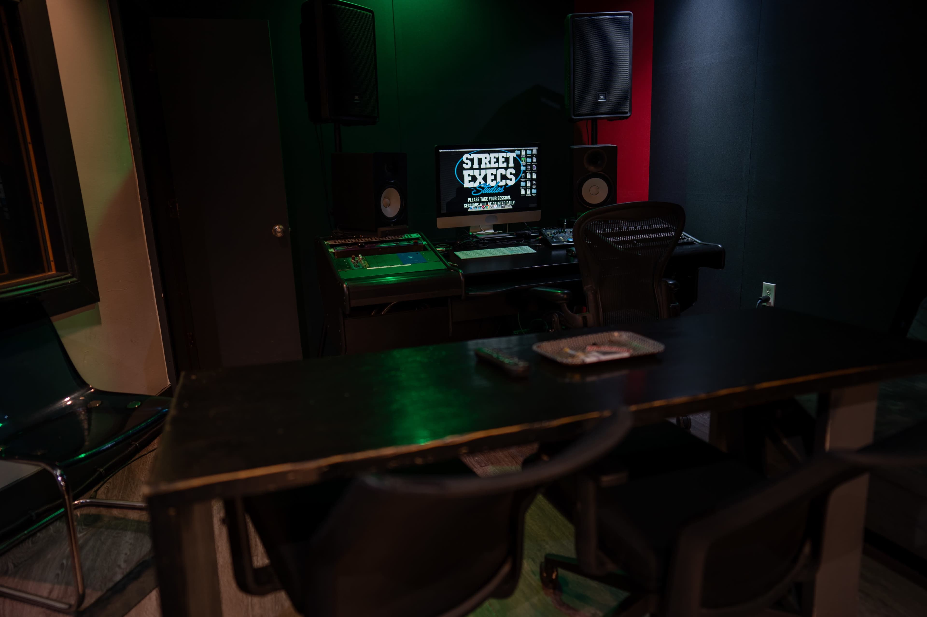 A music production studio featuring a desk with a computer, speakers, and audio equipment, illuminated by green and red lights.