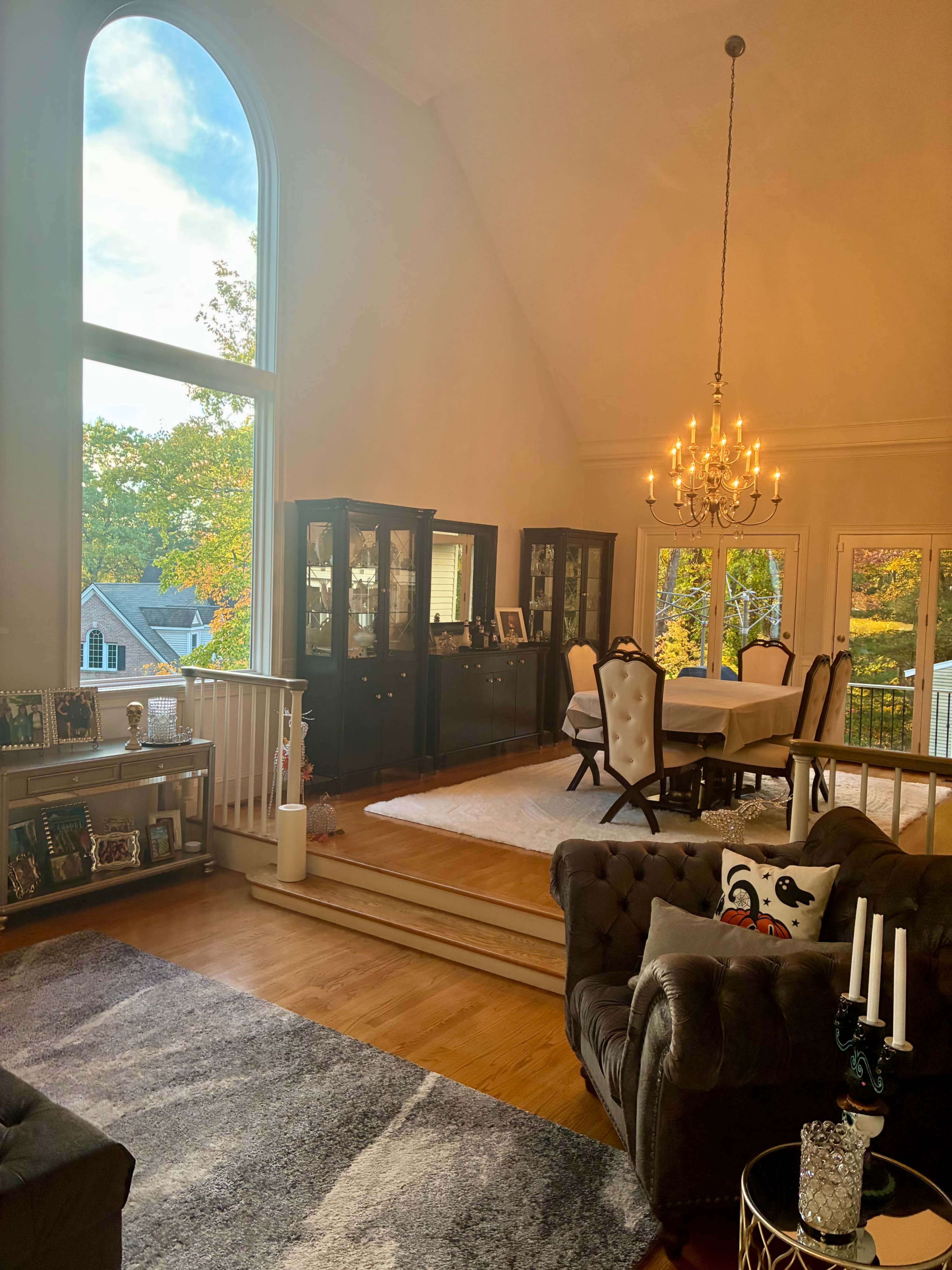 The image shows a spacious living room with a large window letting in natural light, a dining area in the background, and elegant furniture including a chandelier and a plush sofa.
