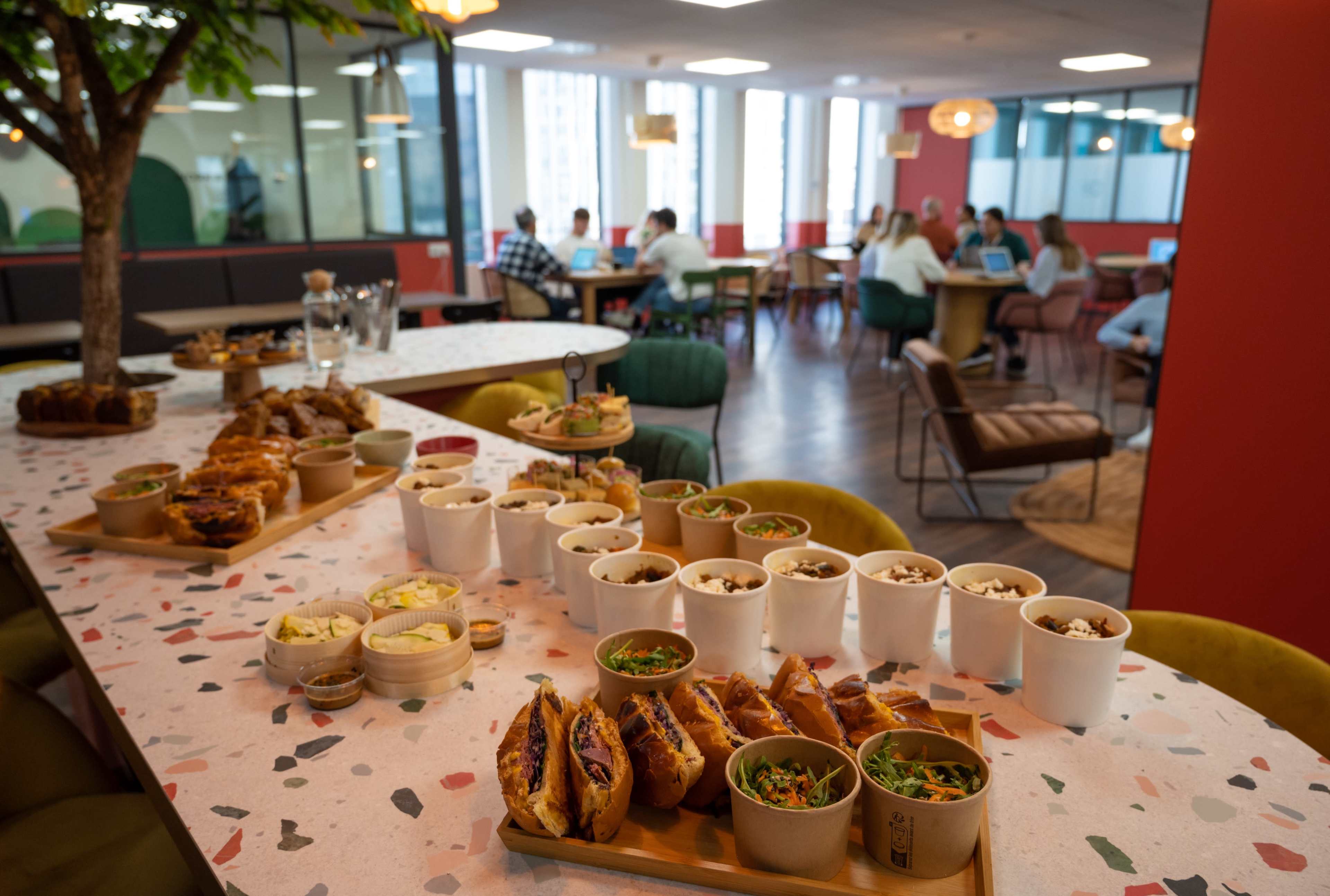 A variety of food items are displayed on a table in an office setting, with people working in the background.