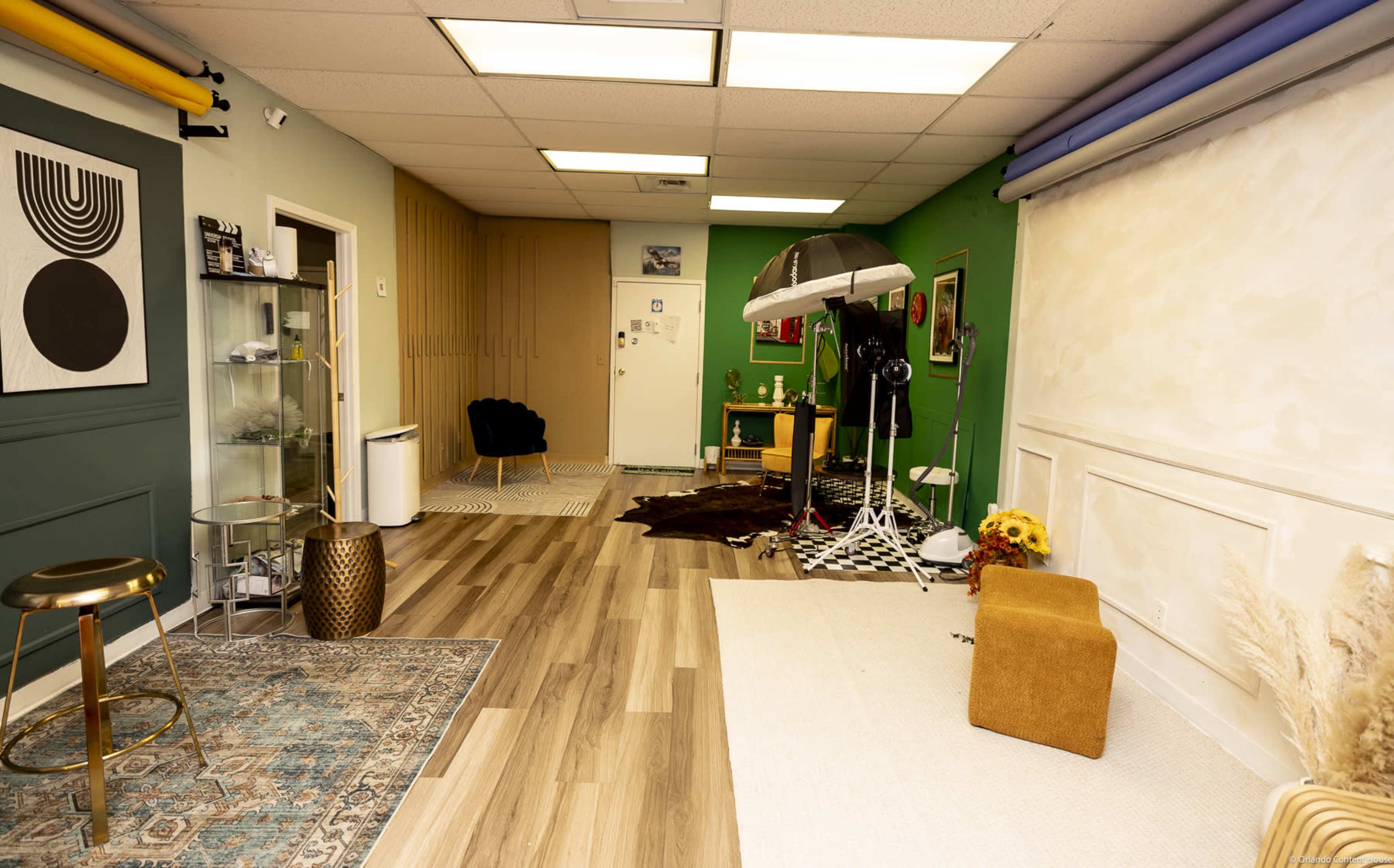 The image shows a spacious room with a mix of green and beige walls, featuring photography equipment, a seating area, and decorative elements like plants and a patterned rug.