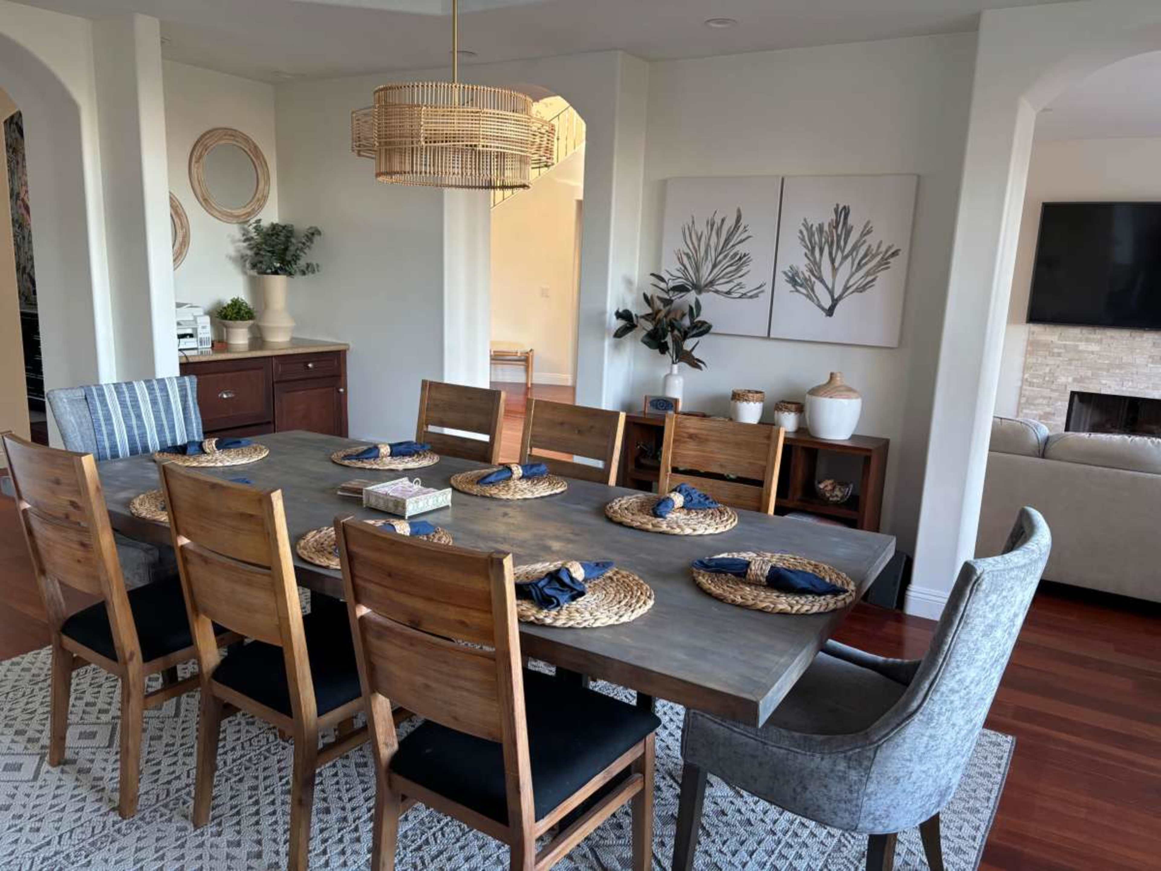 A long wooden dining table set with plates and woven placemats is surrounded by chairs in a well-decorated dining room featuring a light fixture and artwork on the walls.