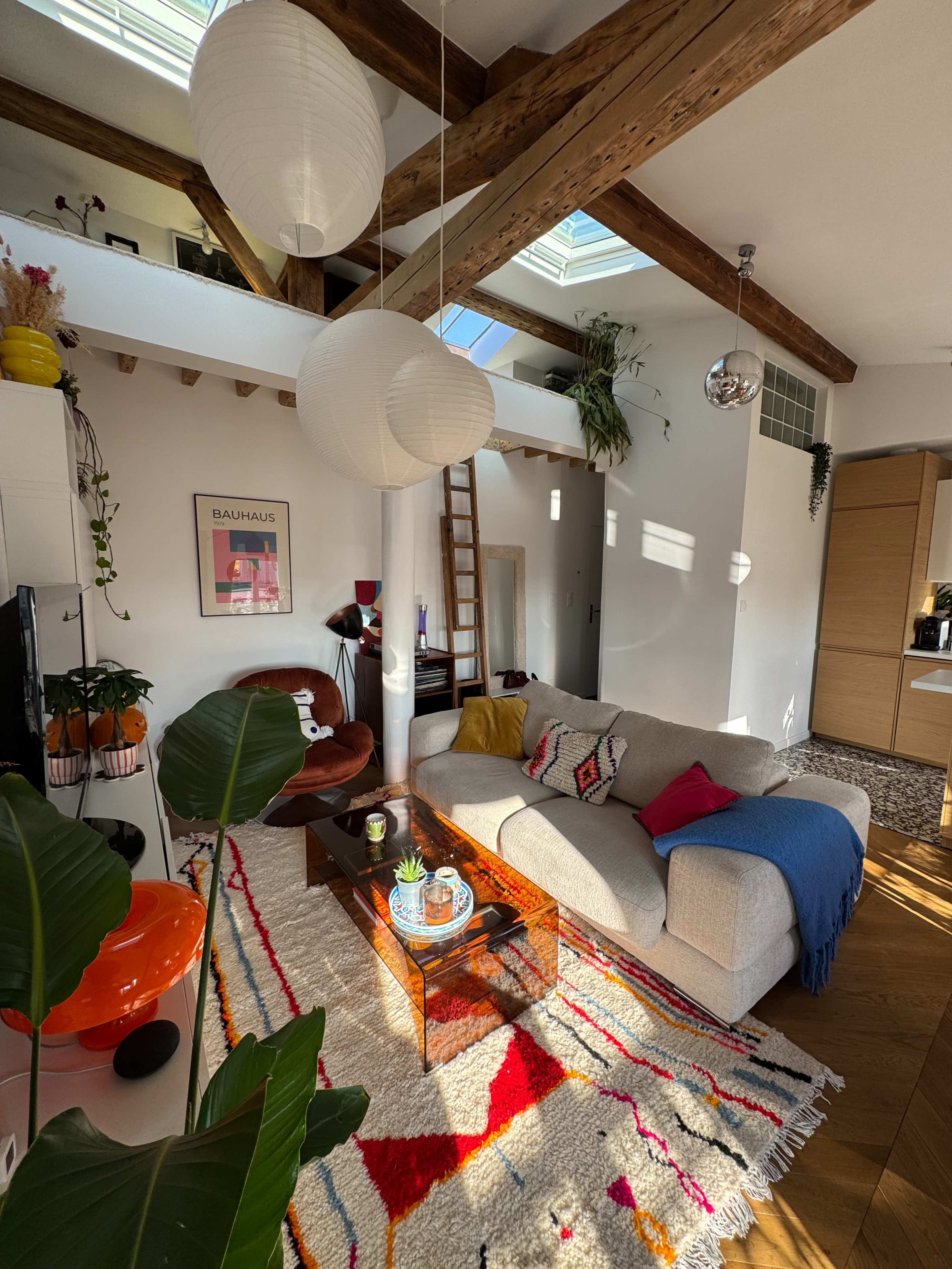 The image shows a cozy living room with a sectional sofa, a glass coffee table, and decorative plants, featuring wooden beams and skylights above.