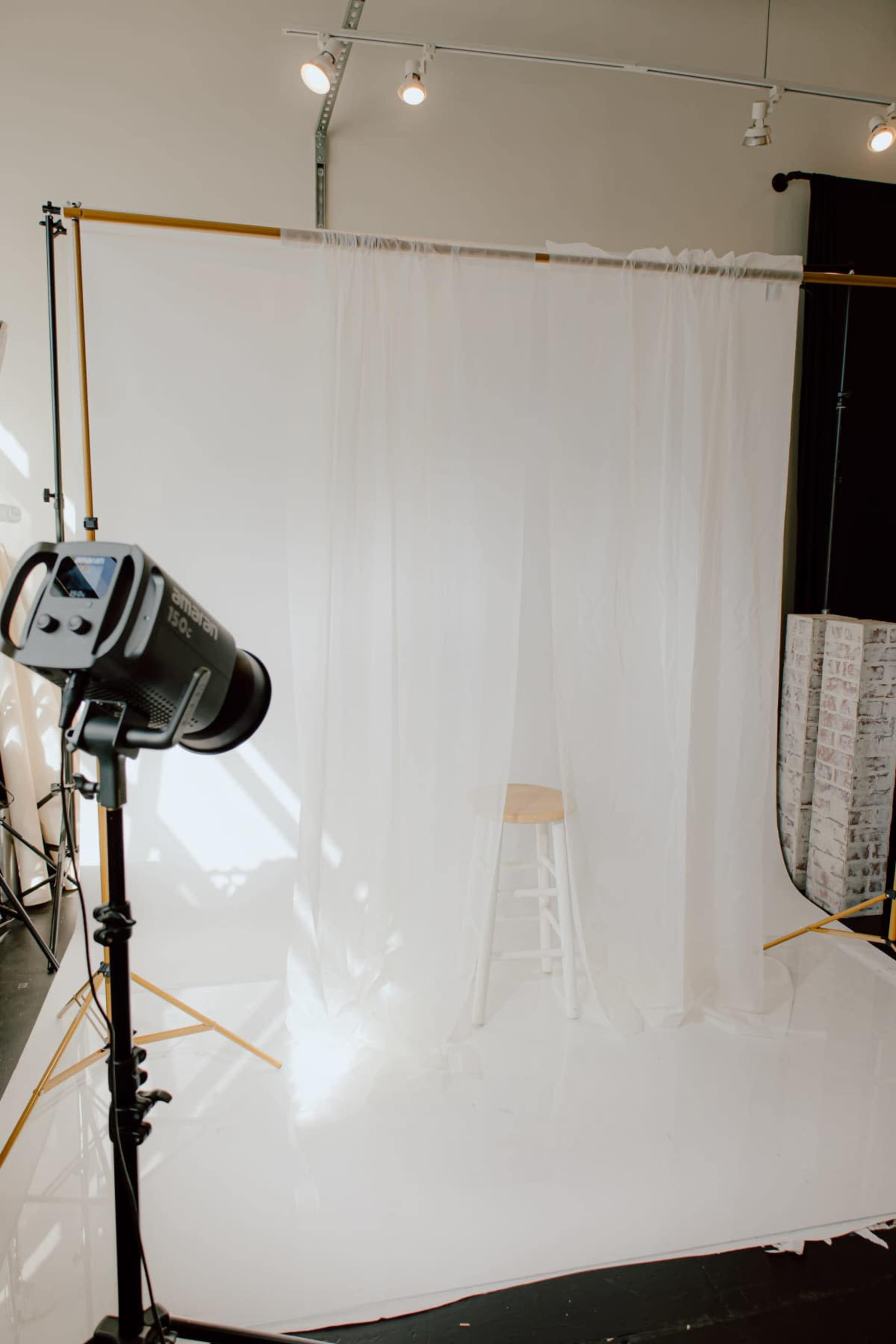 A photography studio is set up with a light source facing a white backdrop draped with sheer fabric and a wooden stool positioned behind it.